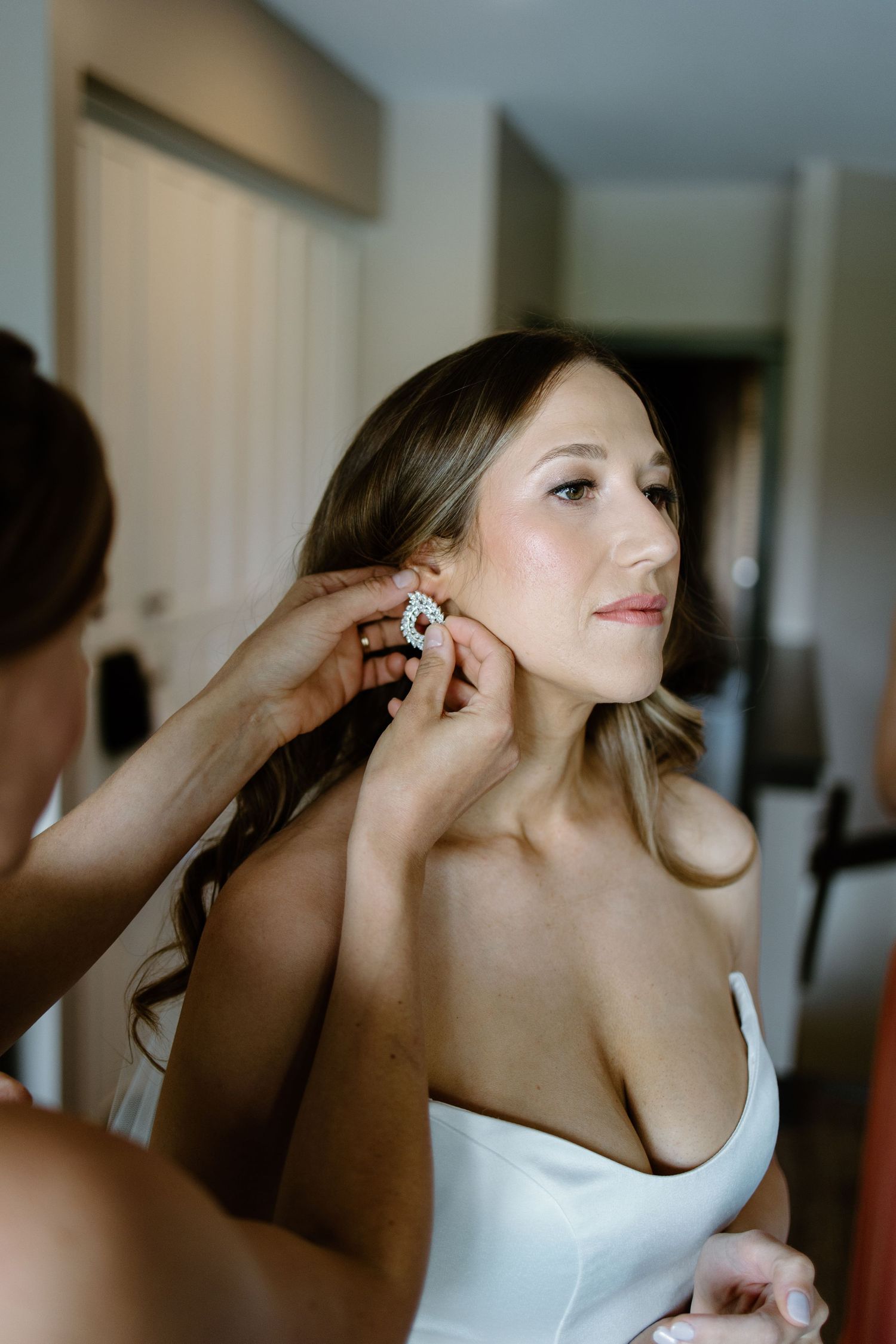 Bride in white dress gets elegant crystal earring put on by bridesmaid while getting ready for wedding.
