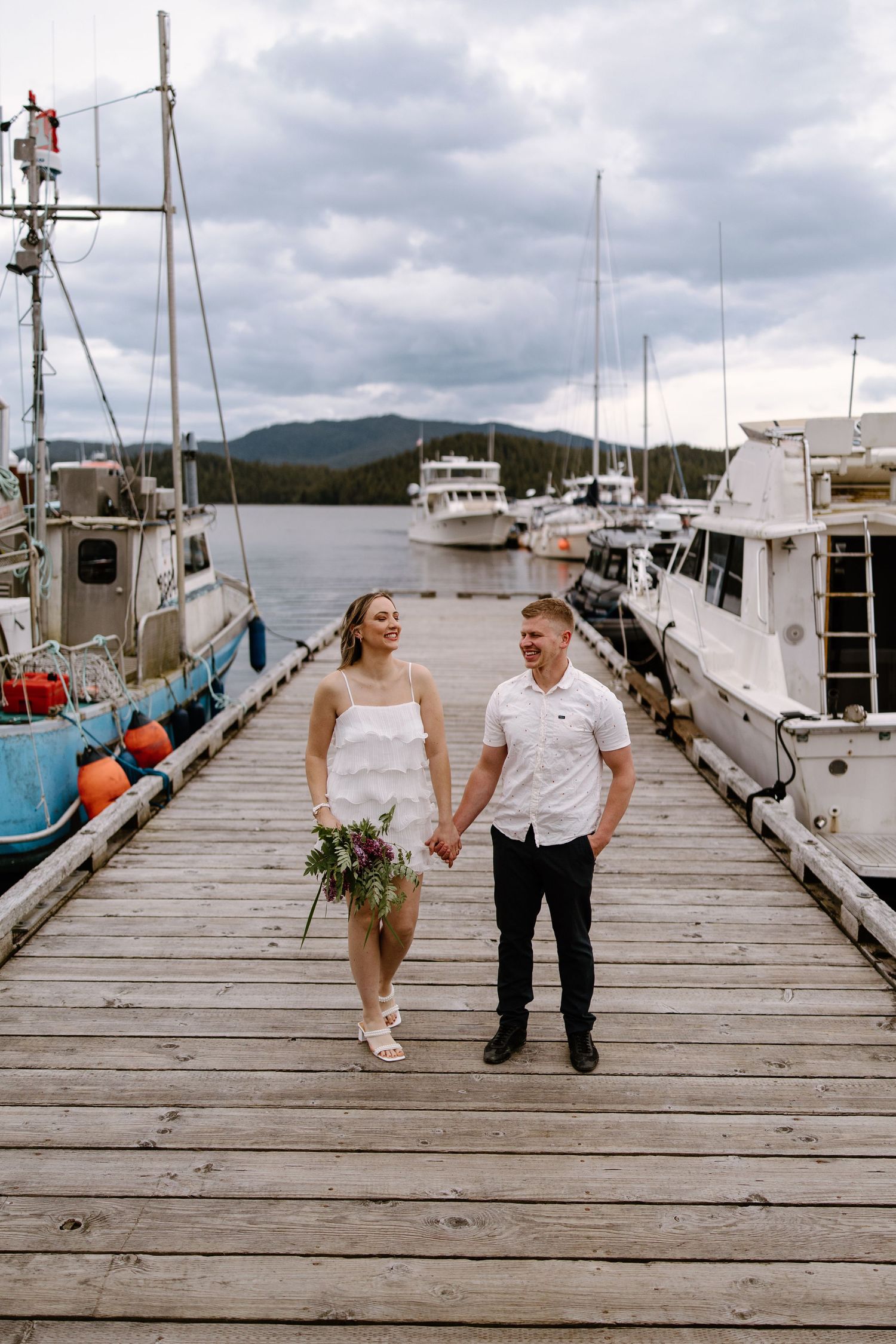 Couple sharing romantic moments while walking along a weathered wooden dock with boats in the marina.