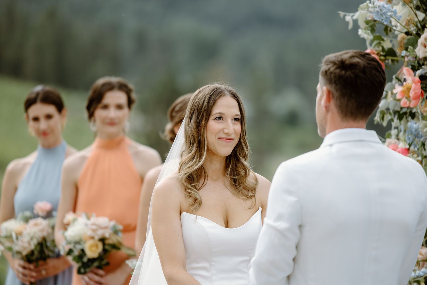 Wedding party in blue and coral dresses holding floral bouquets against scenic mountain backdrop.