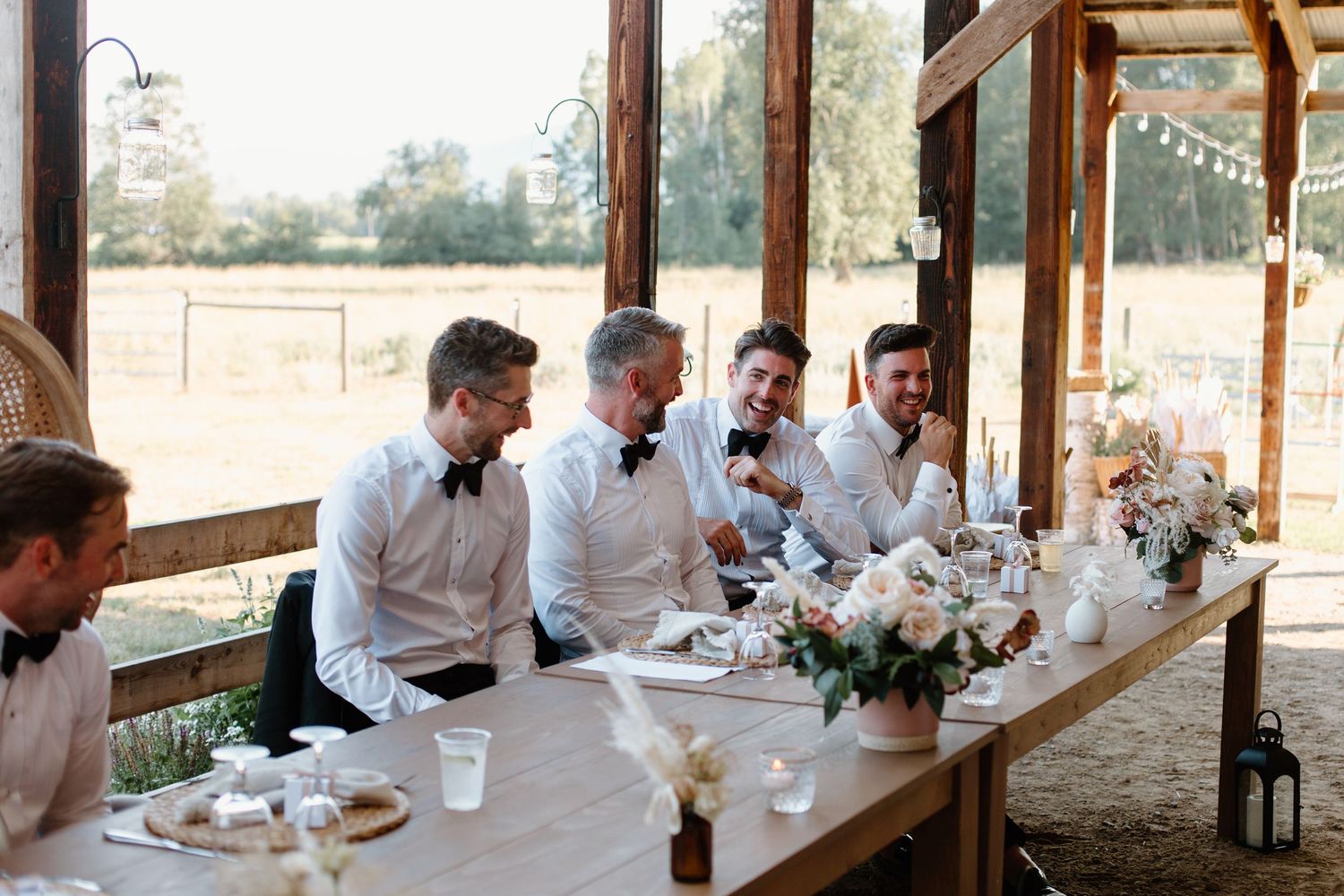 A group in white dress shirts seated at a rustic wooden table during an outdoor wedding reception.