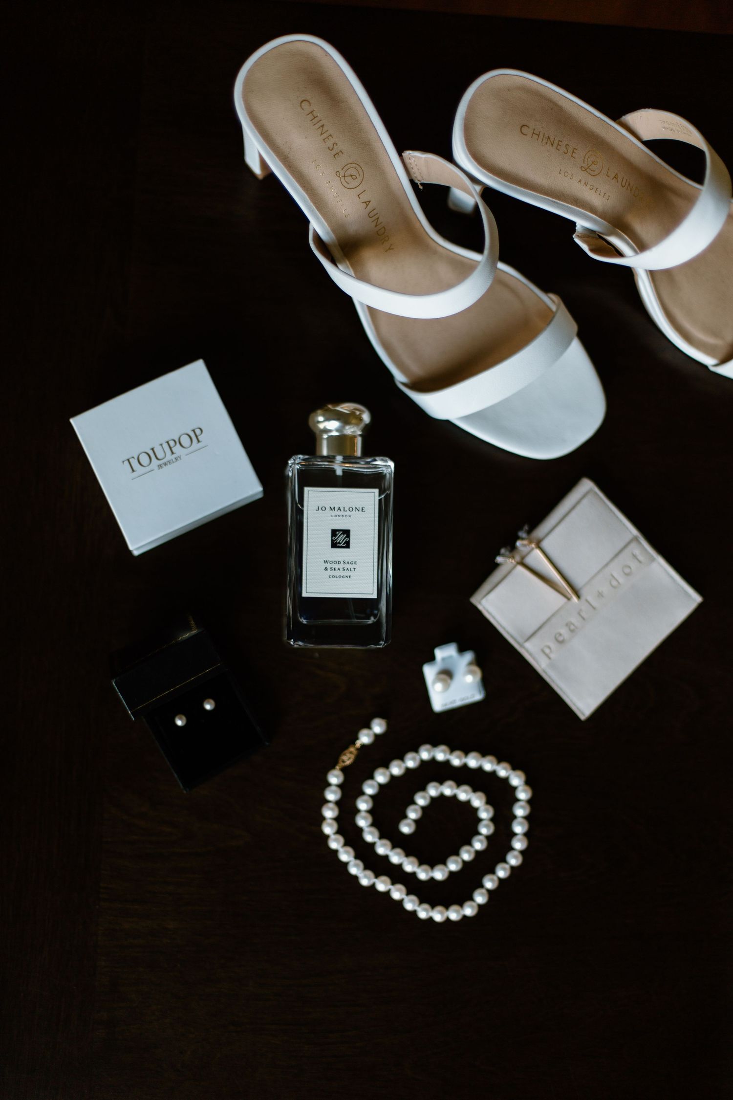 Wedding accessories including white sandals, perfume, pearl necklace and Jo Malone products on dark surface.