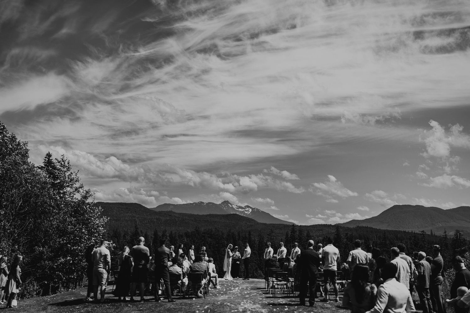 Black and white landscape of wedding guests gathered outdoors with mountains in background.