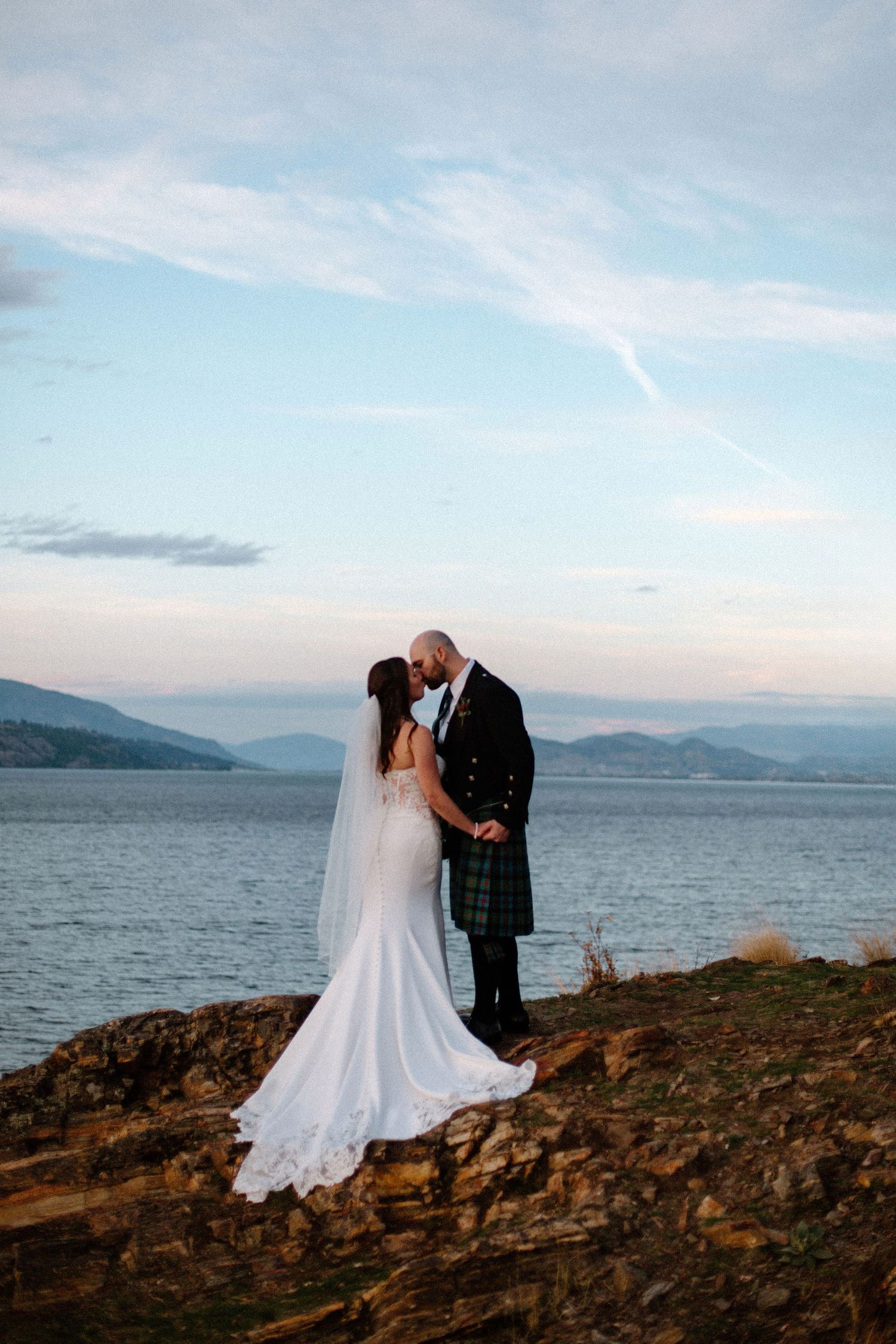 Bride and groom kissing in traditional Scottish kilt attire on rocky shoreline with mountain loch backdrop at sunset.