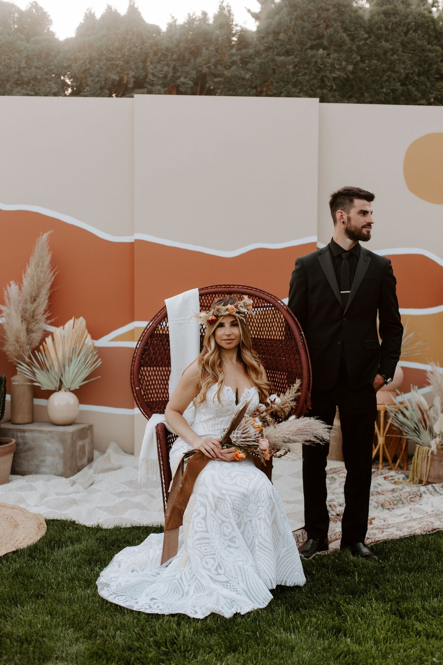Modern boho wedding setup with peacock chair, painted backdrop wall in orange and white, and dried pampas grass decorations.