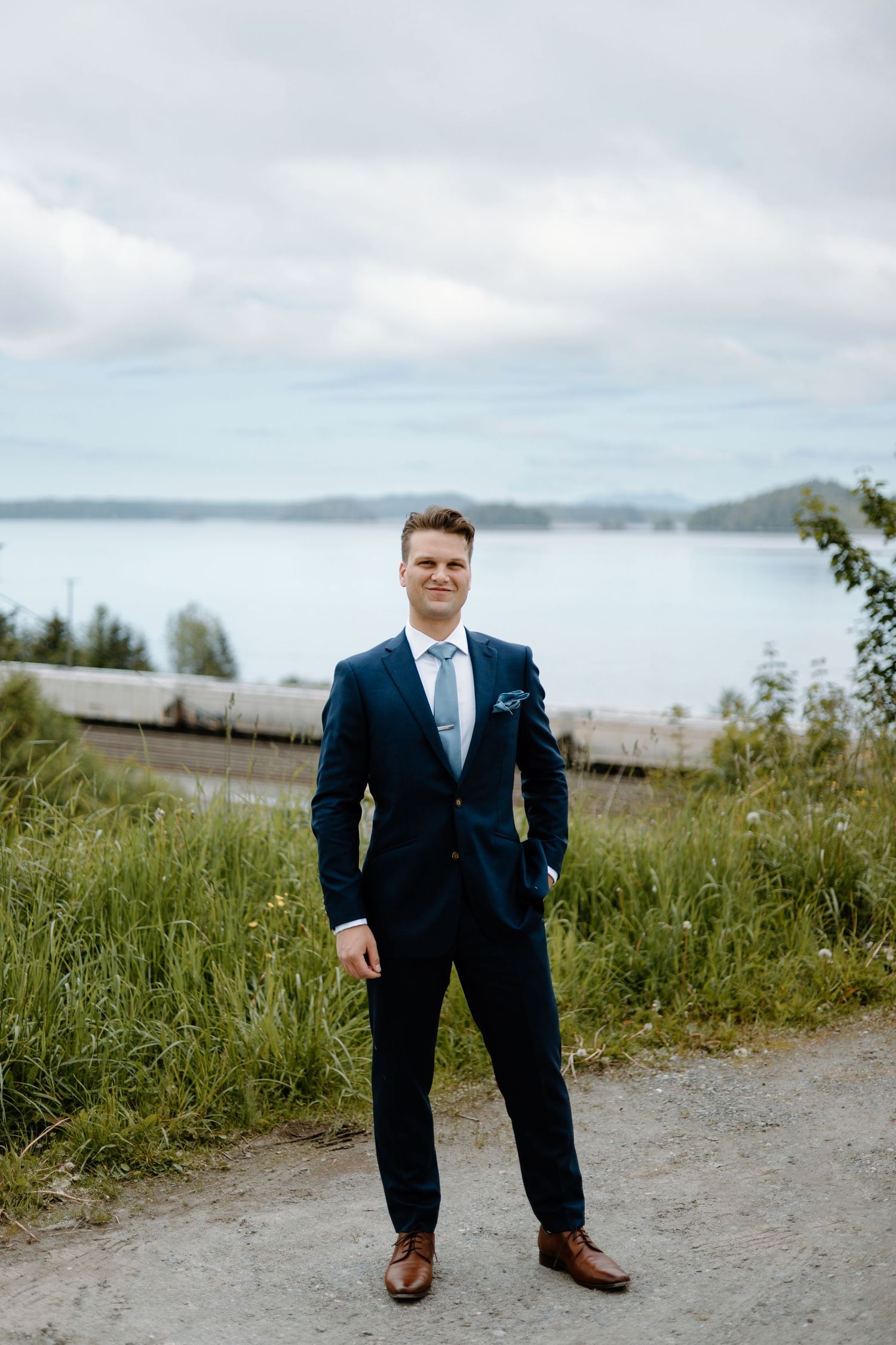 Groom in navy suit with light blue tie stands on gravel path with lush greenery and water in background.