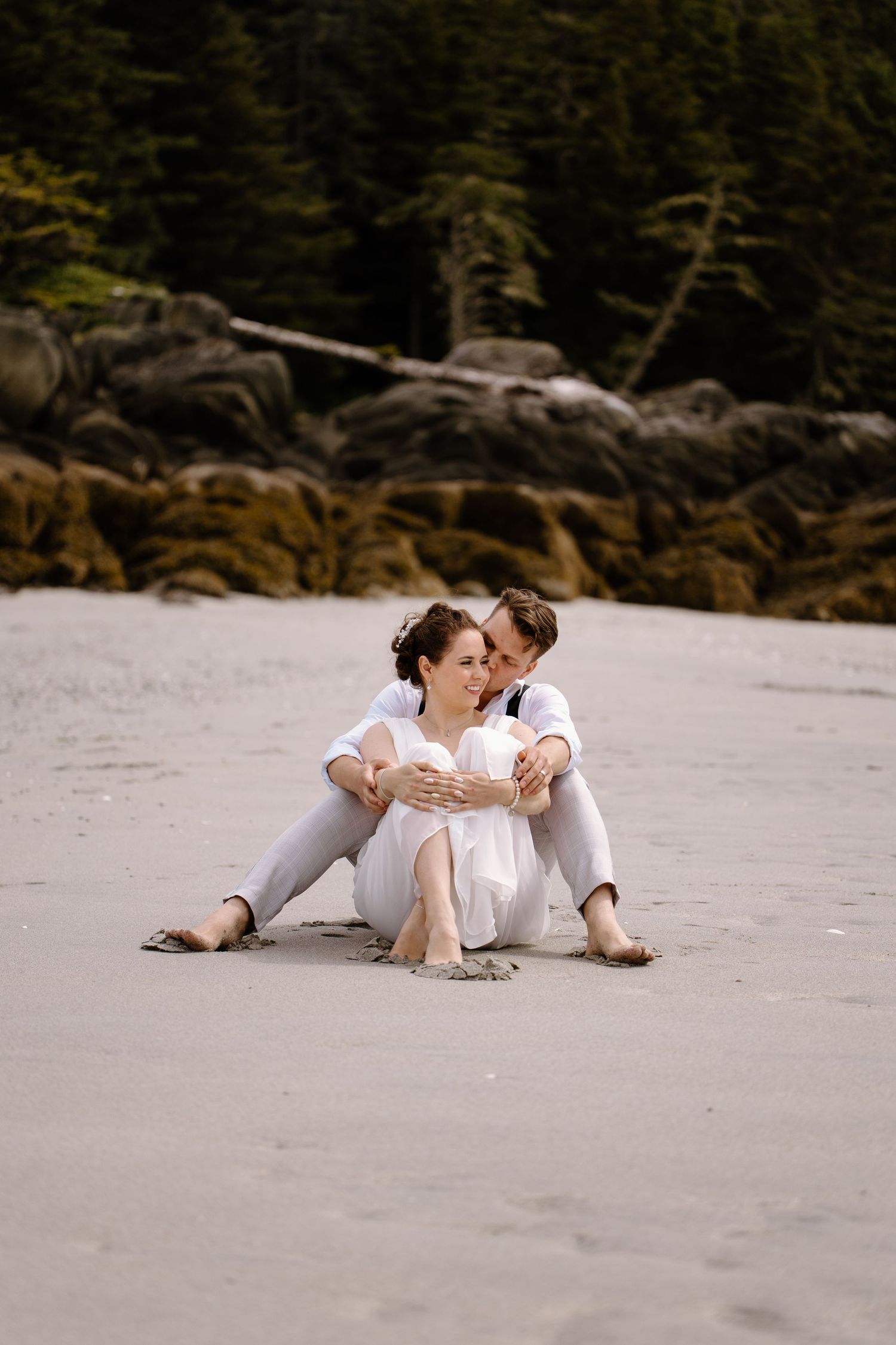 A couple in white outfits reclines together on a sandy beach with evergreen trees in the background.