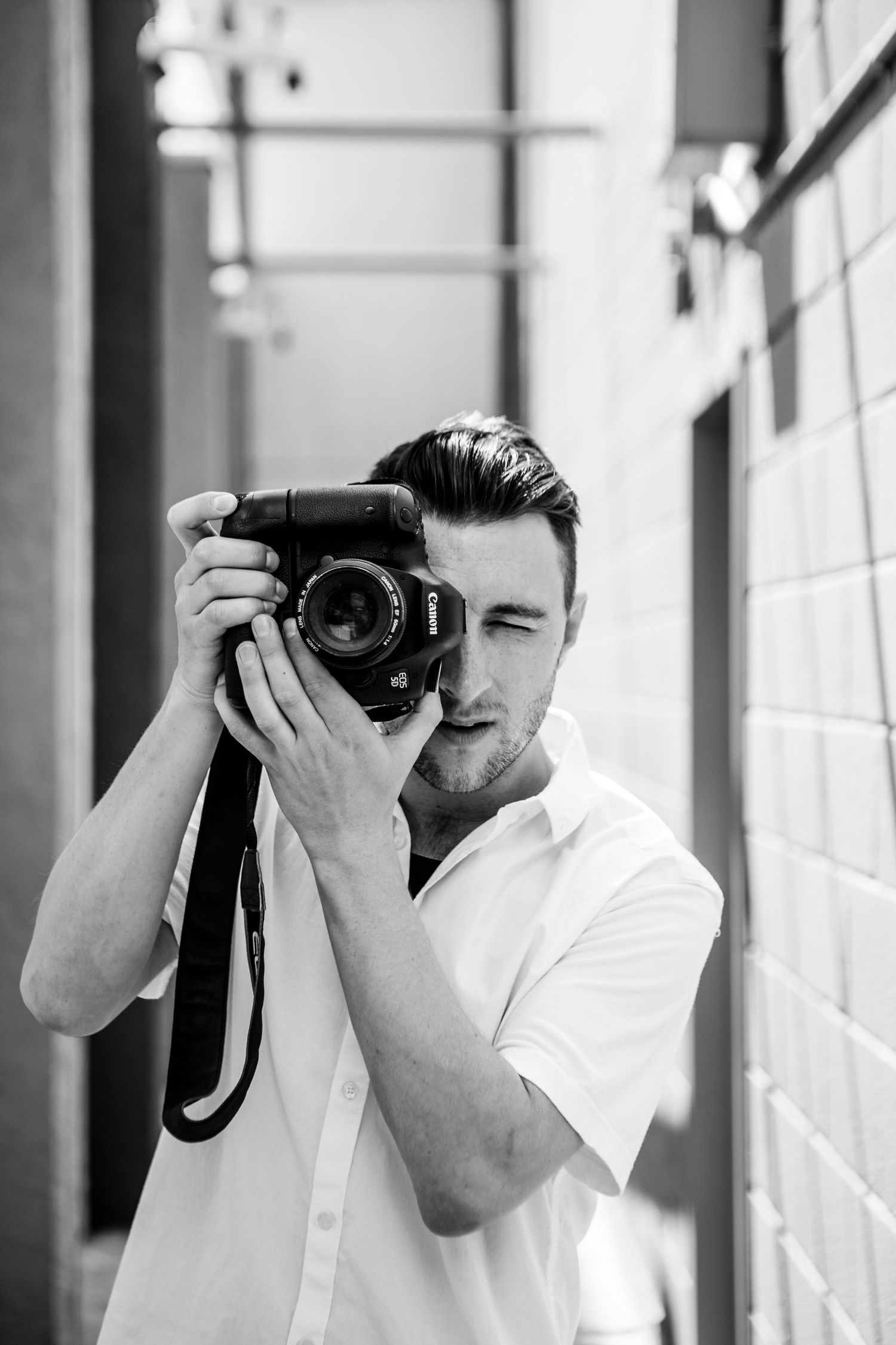 Photographer in white shirt capturing photos with professional camera against light wall.