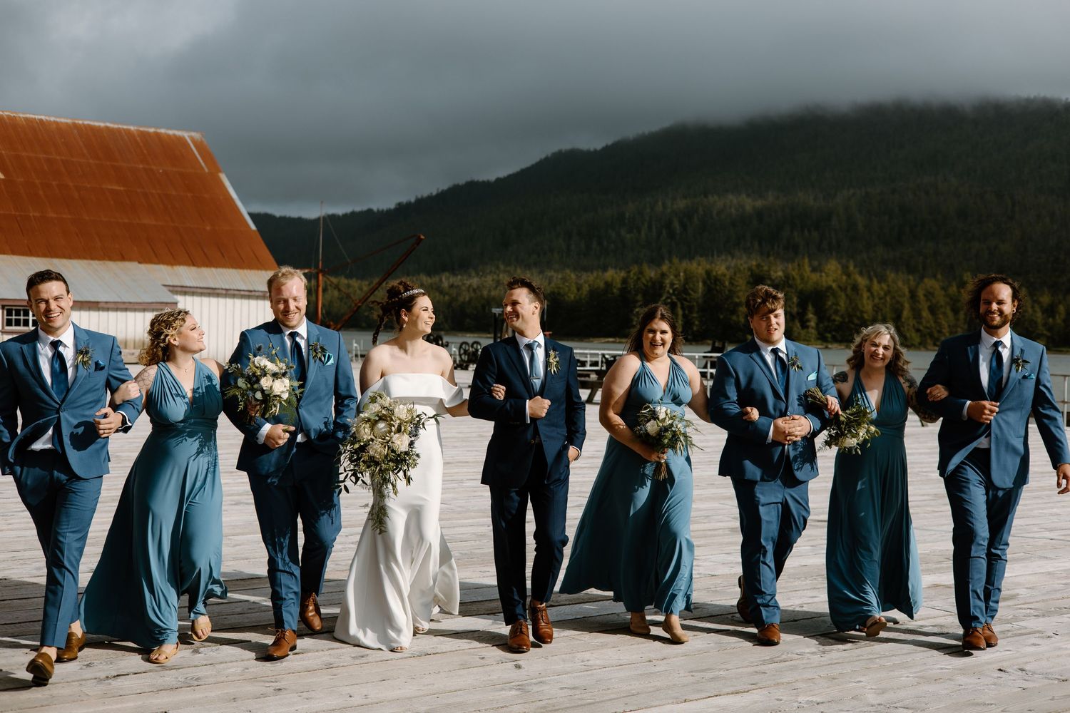 Wedding party walks in sync wearing coordinated dusty blue attire against a mountain backdrop.