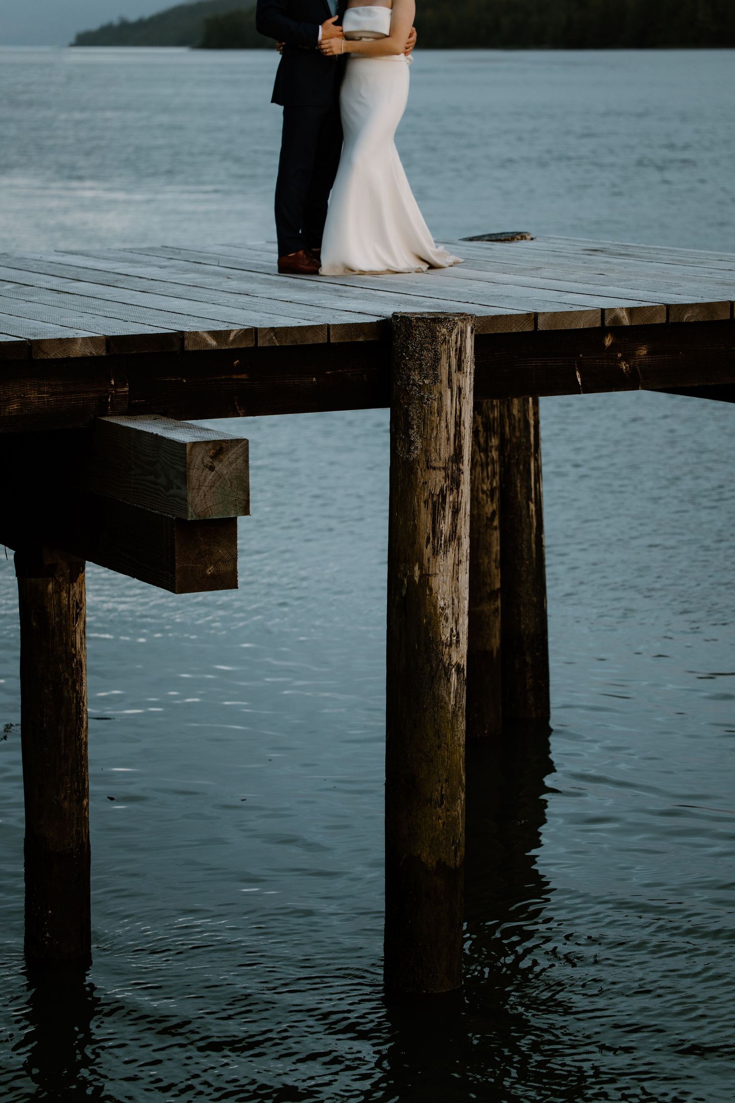 A couple in wedding attire stands together on a wooden dock overlooking calm waters.