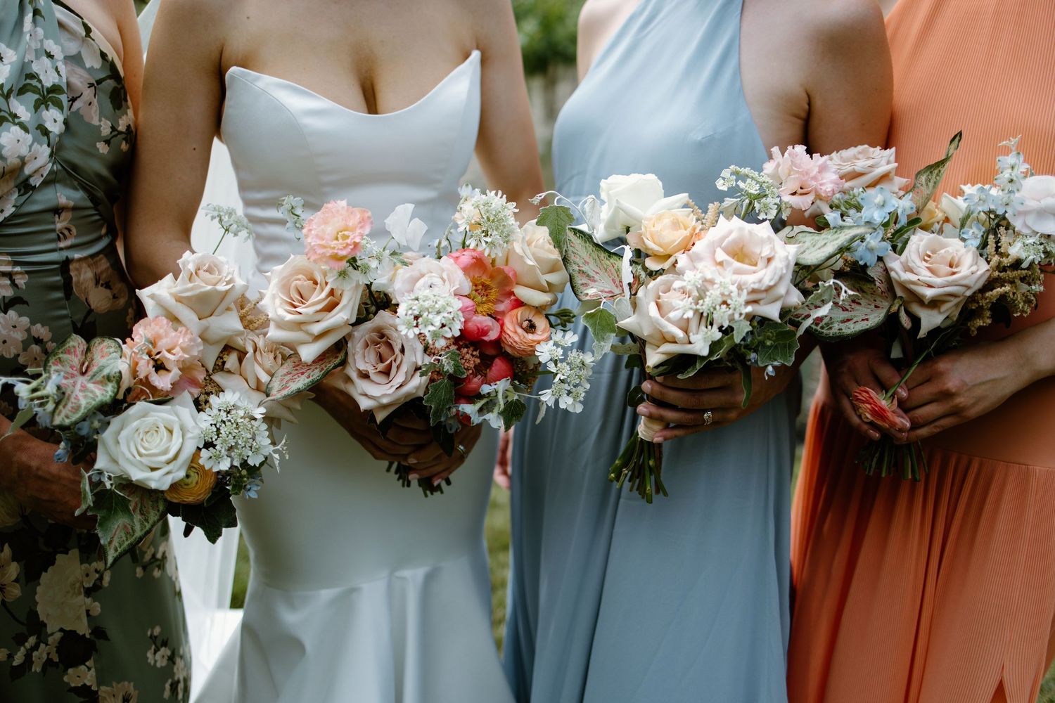 Row of bridesmaids holding blush and coral flower bouquets at outdoor wedding.