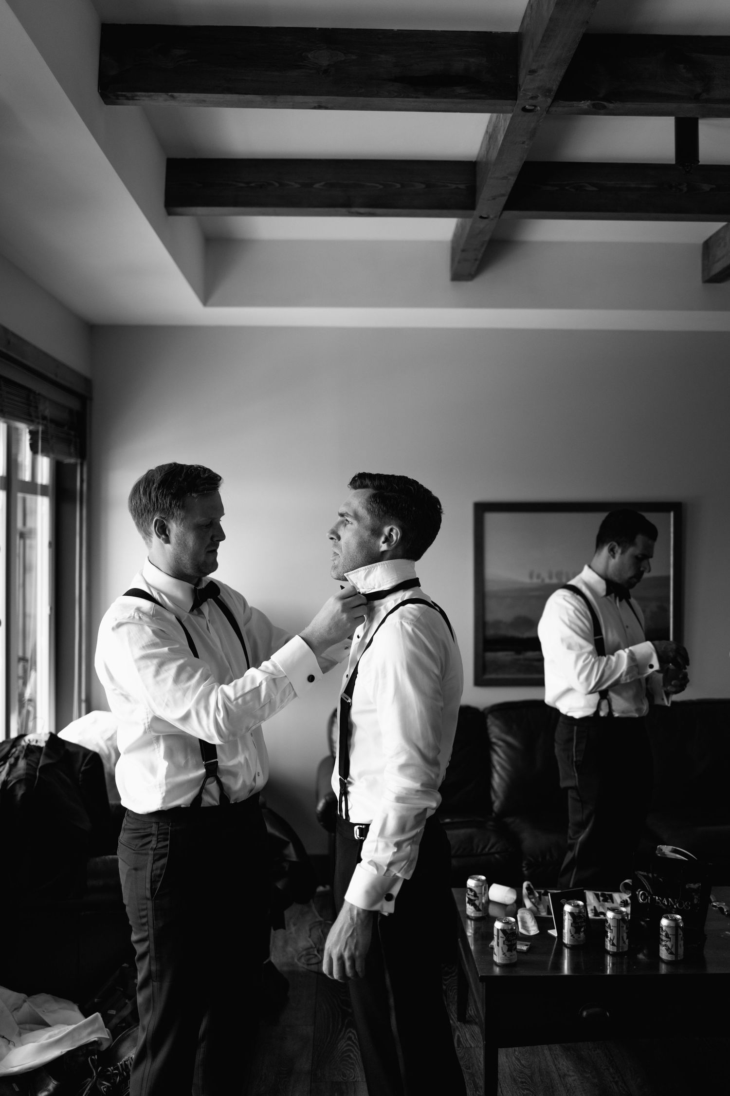 Black and white photo of groomsmen helping each other with bow ties and suspenders while getting ready for a wedding.