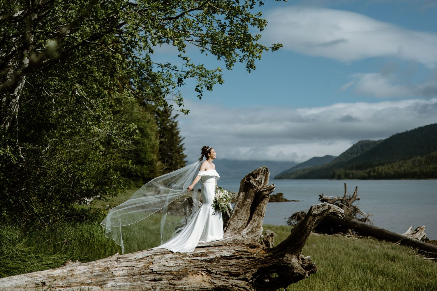 Bride in white gown with flowing veil stands on driftwood by a mountain lake surrounded by lush green trees.
