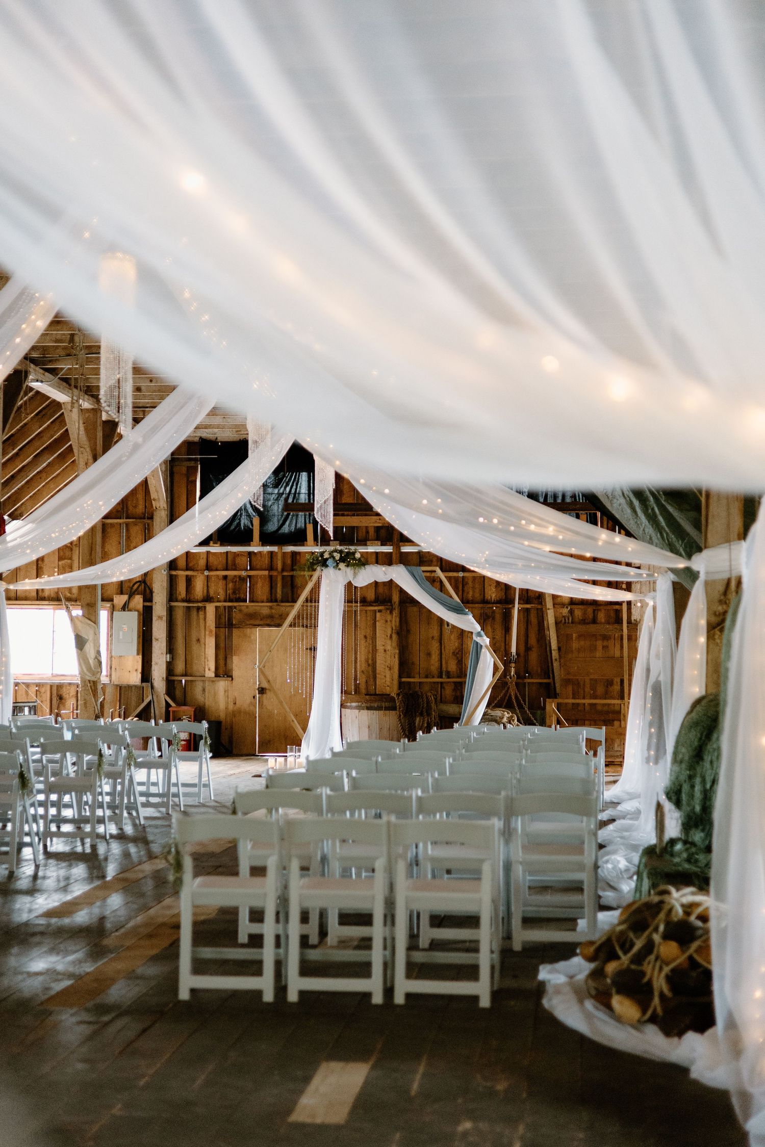 Rustic barn wedding venue with white draped fabric ceiling, string lights, and rows of white folding chairs for ceremony.
