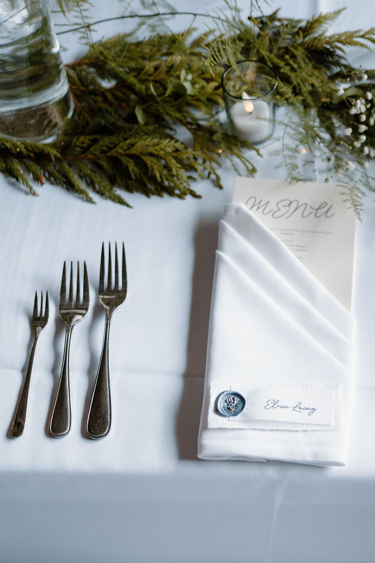 Place setting with silver forks and menu card decorated with evergreen branches and candlelight.