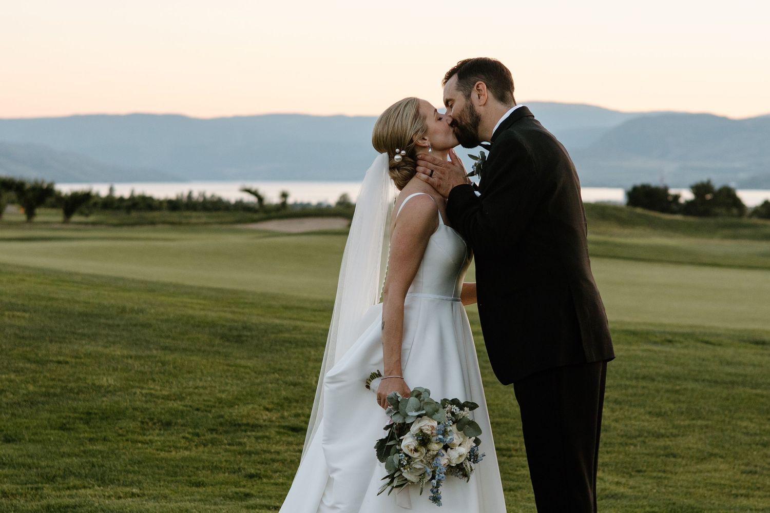 Bride and groom share an intimate moment on a golf course with mountain and lake views at sunset.