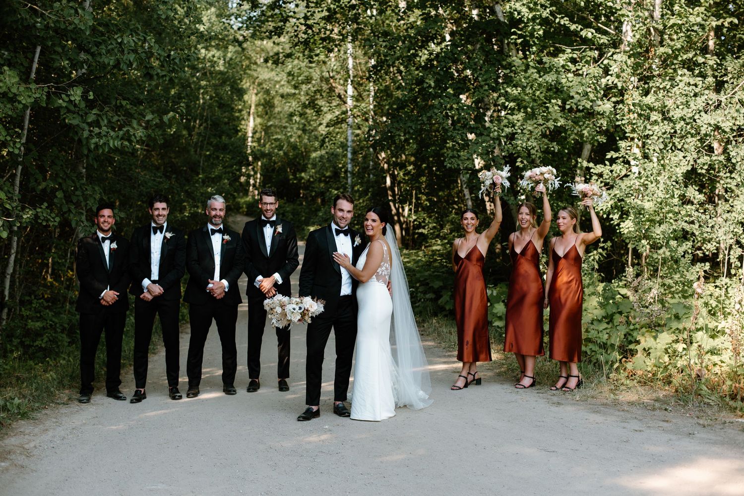 Wedding party in formal attire gathered outdoors against natural backdrop.