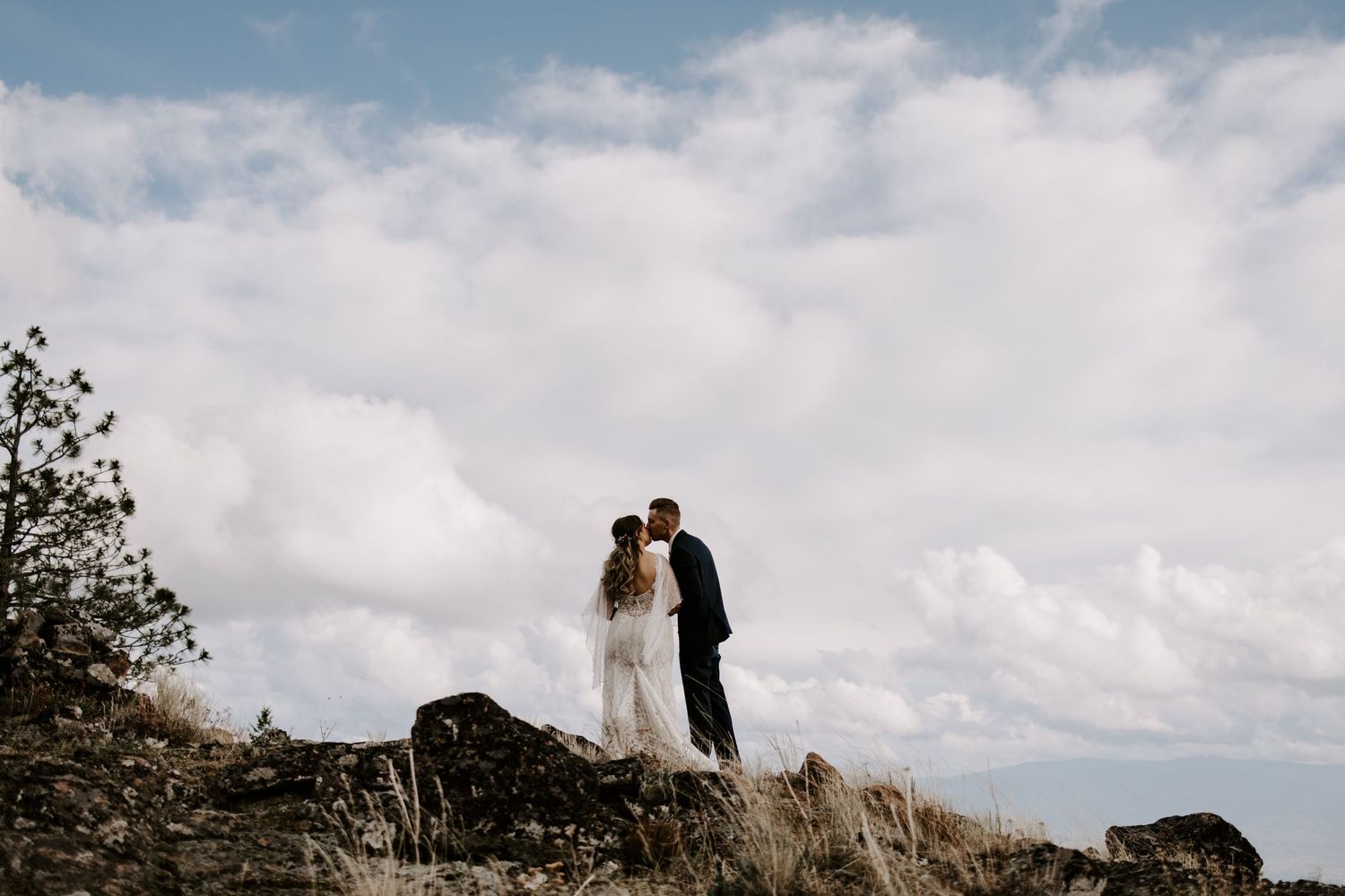 A couple stands in silhouette against cloudy skies on a rocky hilltop.