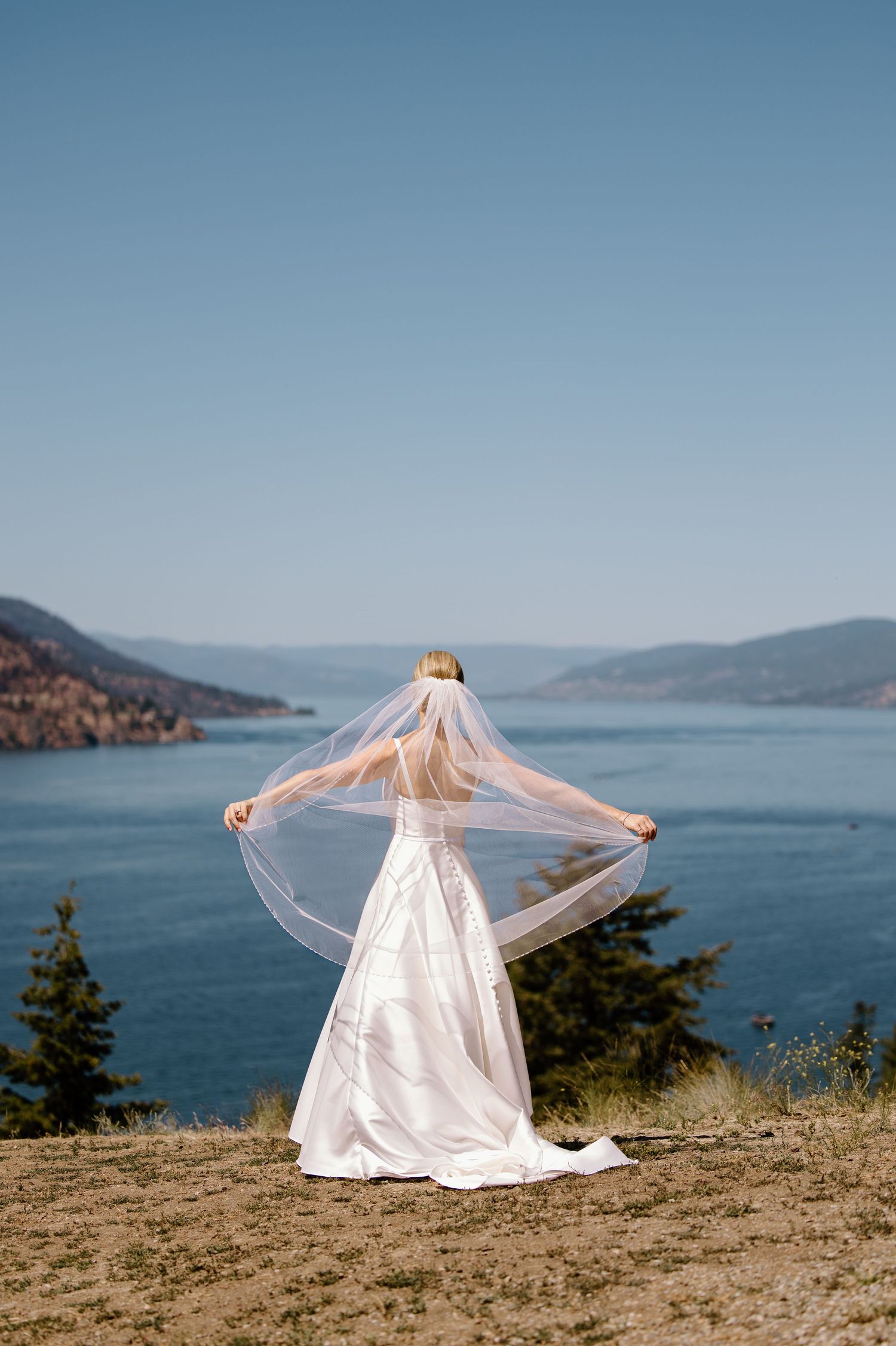 A bride in a flowing white dress and veil stands on a cliffside overlooking a scenic mountain lake on a clear day.