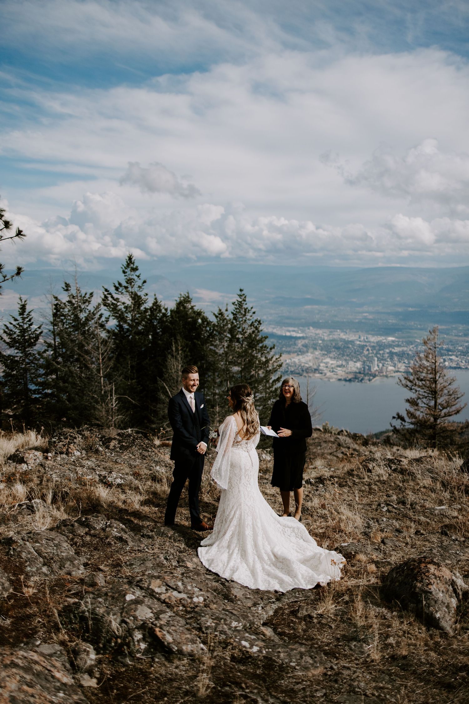 Wedding ceremony group stands on rocky mountain peak overlooking city and water views below.