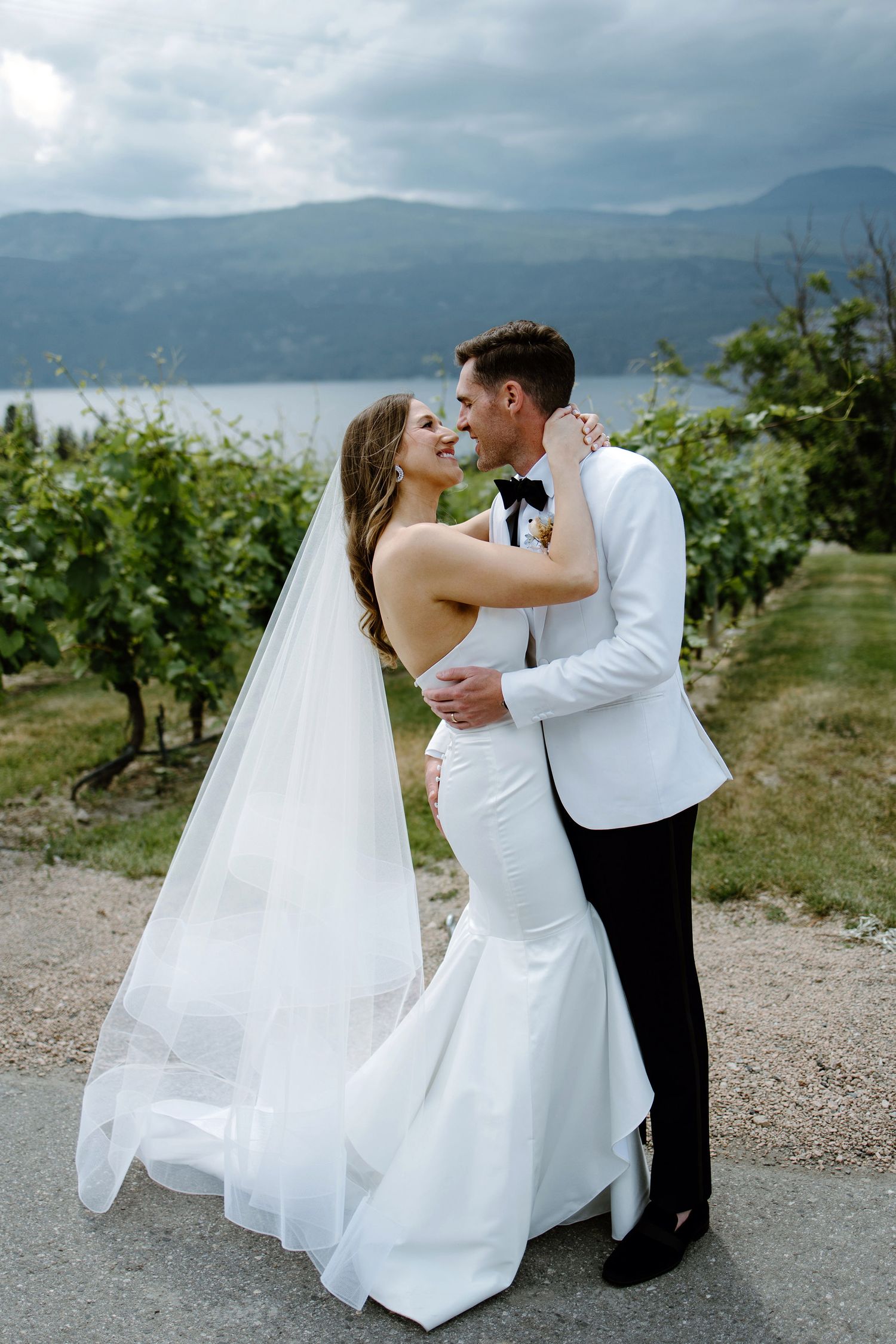 A couple embraces in wedding attire at a vineyard overlooking a scenic mountain lake.