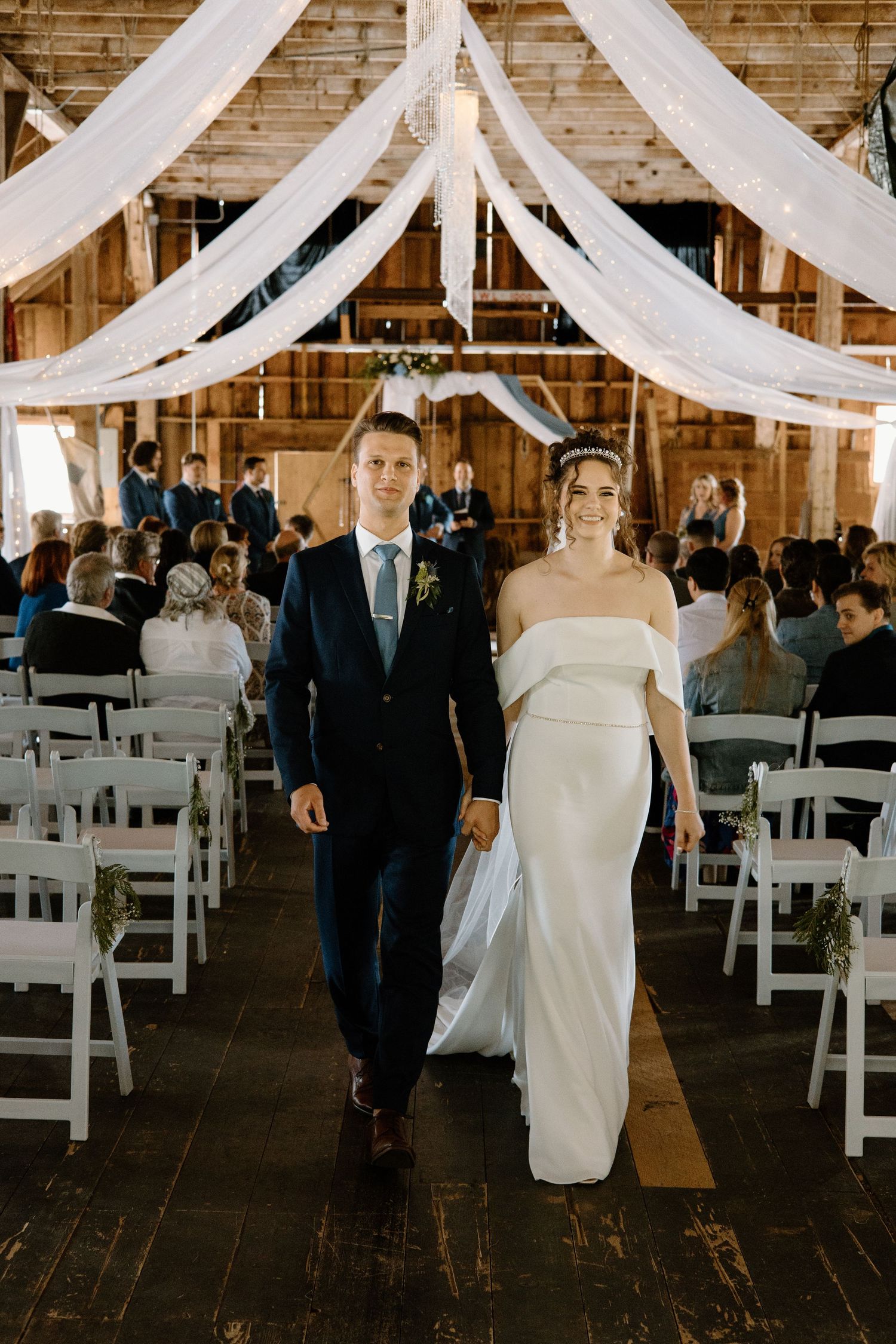 Bride and groom walk down the aisle of a rustic barn wedding venue with white draping and seated guests.