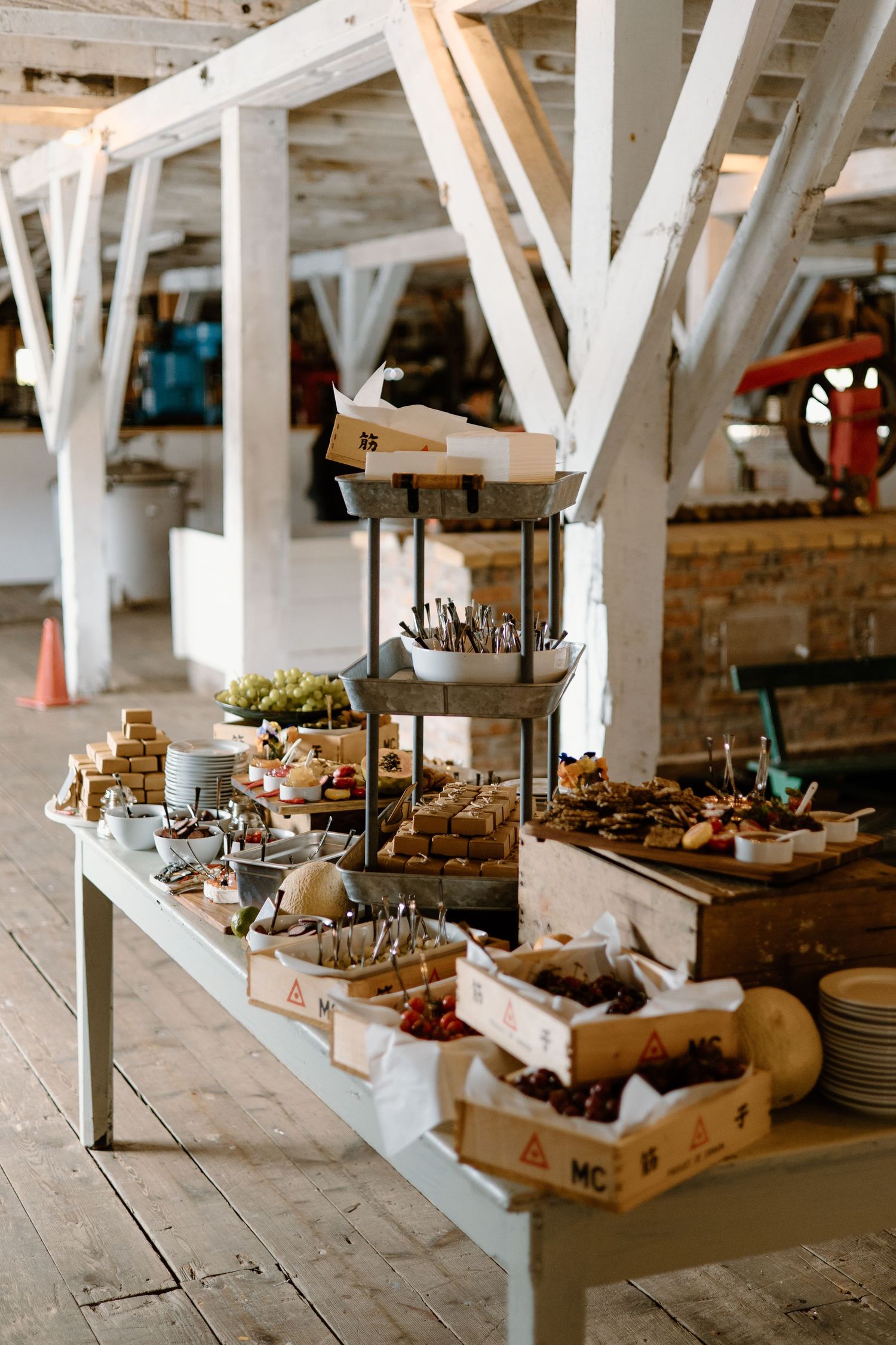 Rustic wedding dessert and appetizer display table with tiered serving platters under white beams.