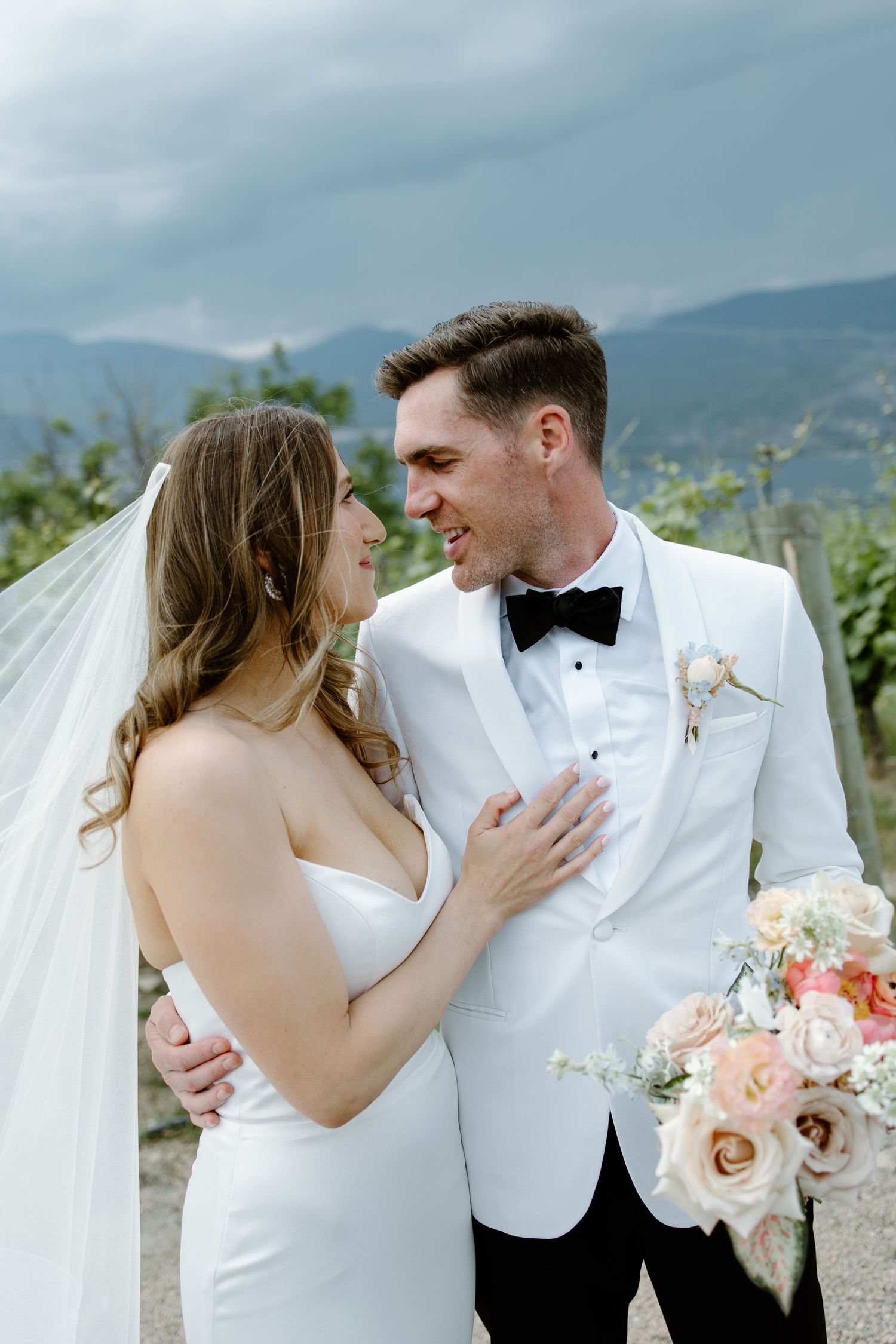 A bride and groom share a romantic moment in a vineyard, with mountains in the background, as she holds a pastel bouquet.