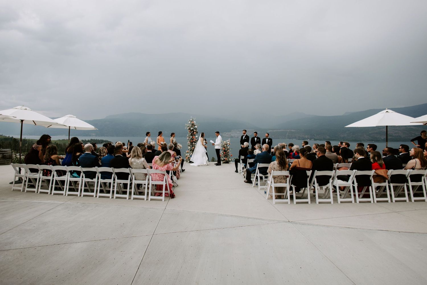 Outdoor wedding ceremony on a rooftop terrace overlooking misty mountains and a lake, with guests seated in white chairs.
