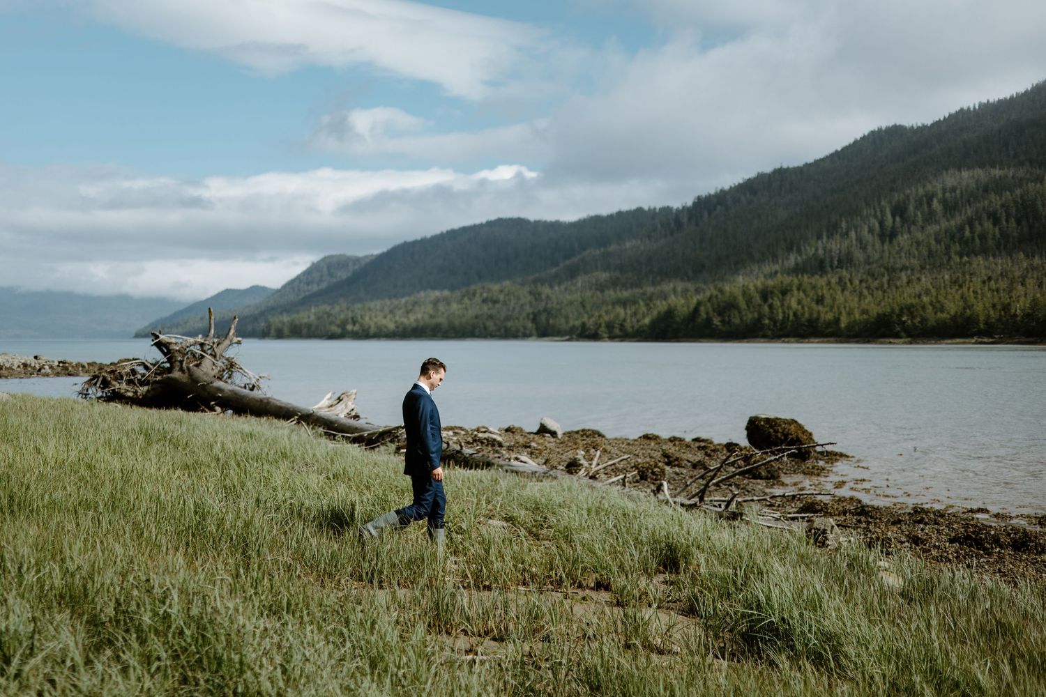 A groom in a navy suit walks along a scenic shoreline with mountains and lush greenery in the background.