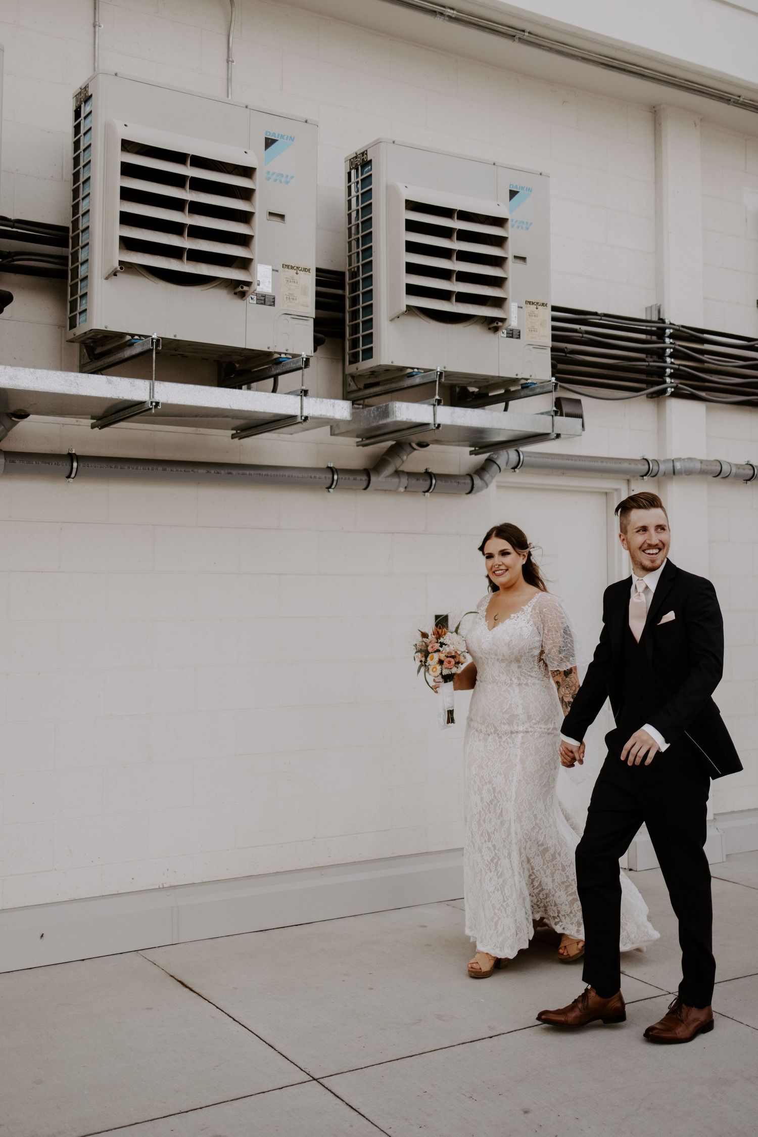 A bride in a lace dress and groom in black suit stand against an industrial wall with ventilation units.