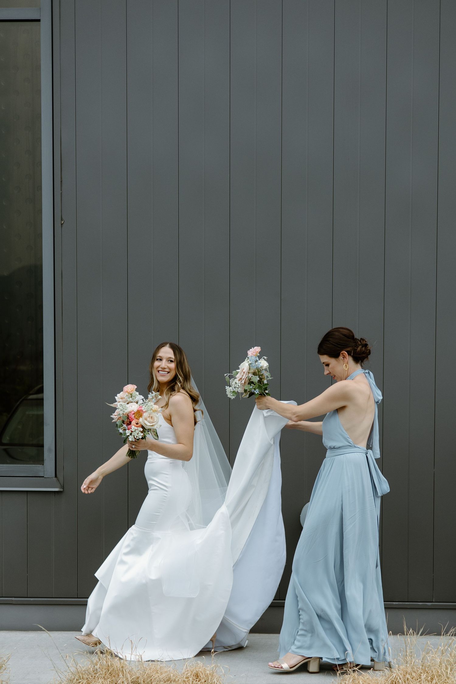 Light blue bridesmaid dresses complement a flowing white wedding gown against a grey exterior wall.