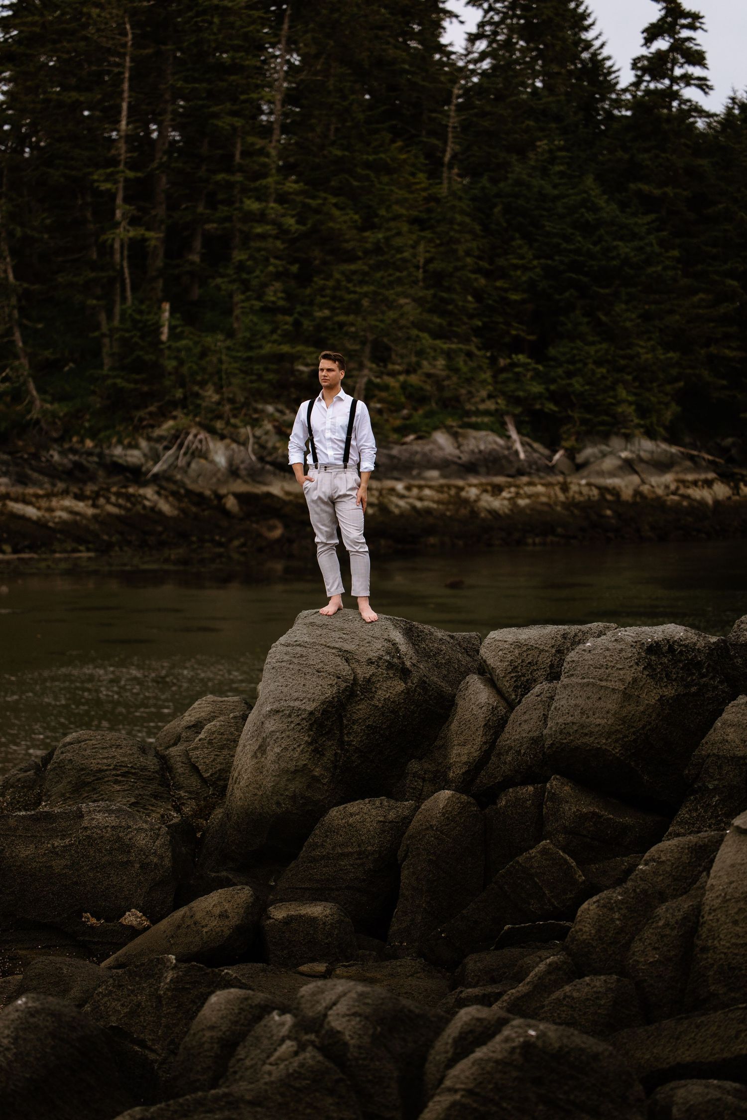 Person in white outfit stands contemplatively on rocky coastline with evergreen forest backdrop at dusk.