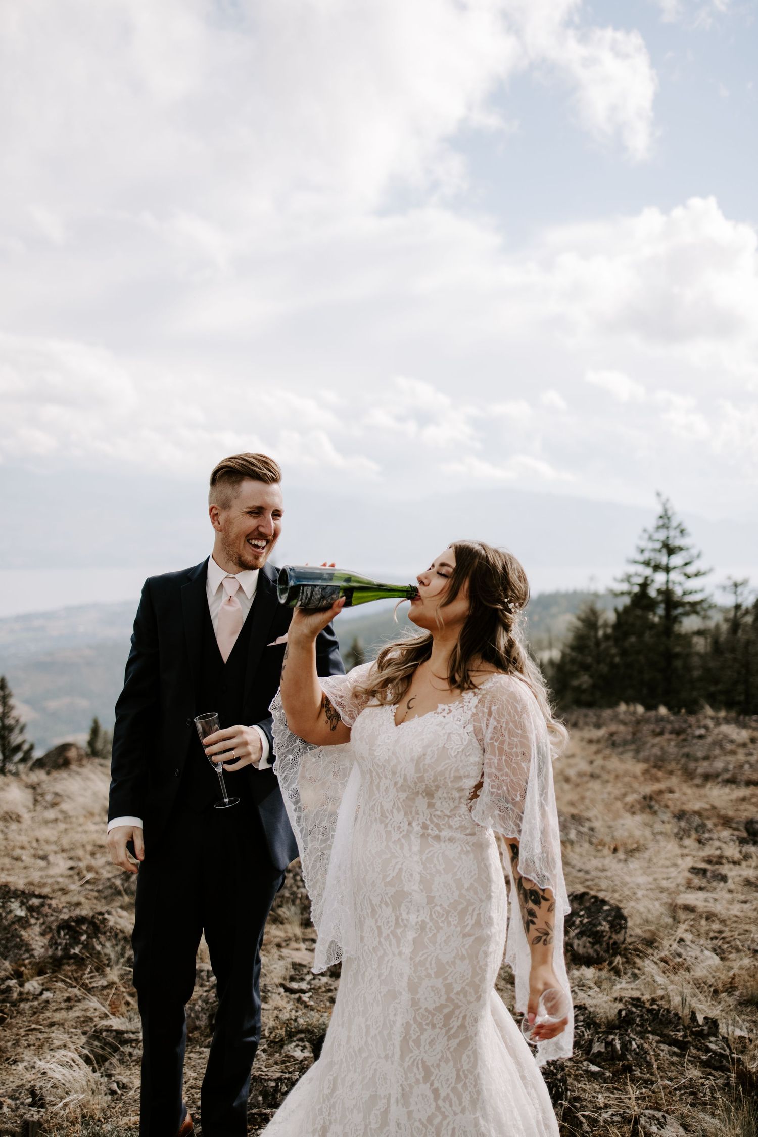 Newlyweds share a joyful moment together on a mountain ridge with dramatic cloudy skies in the background.