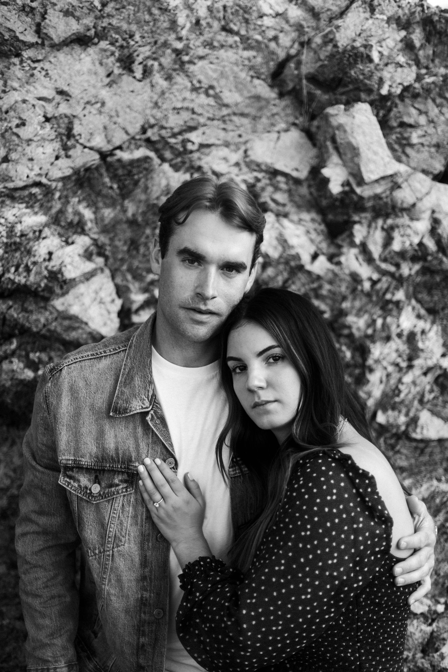 A romantic black and white portrait of a couple posed against a textured rock wall.