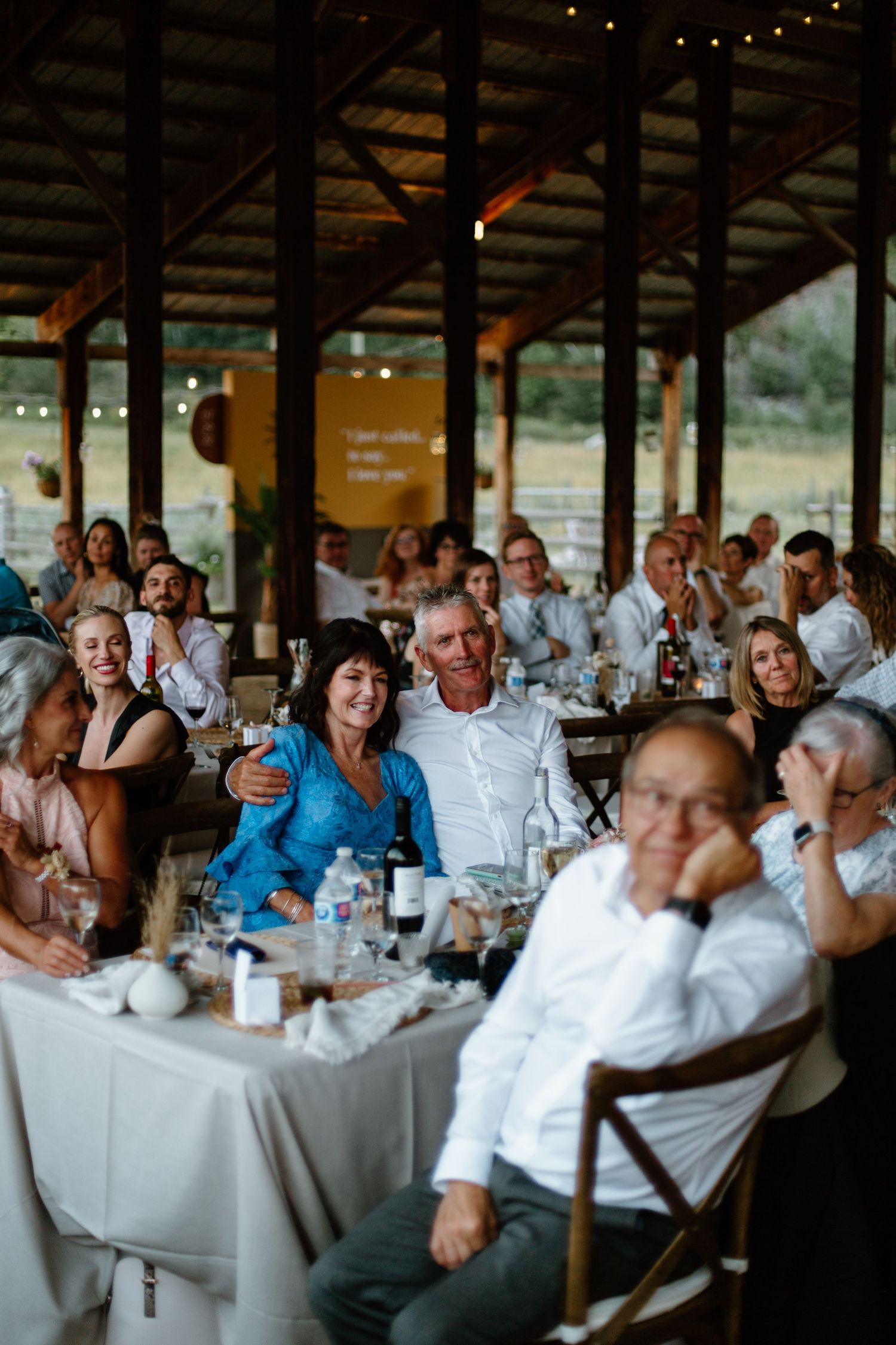 Wedding guests seated at long tables with white linens enjoy a reception dinner under string lights at an outdoor venue.