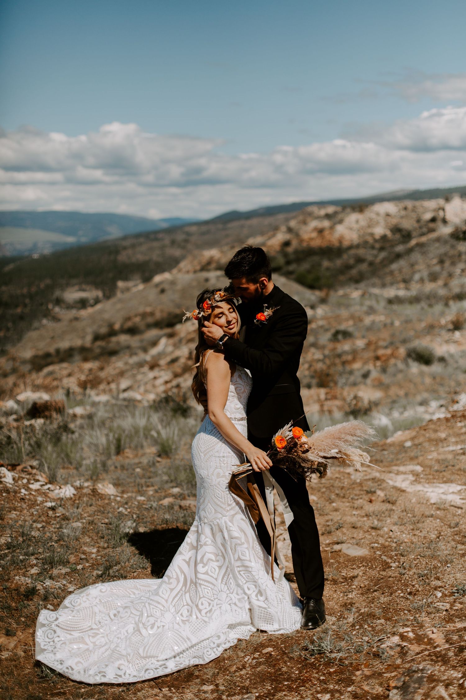 Wedding couple shares an intimate moment on a scenic mountain ridge under blue skies.