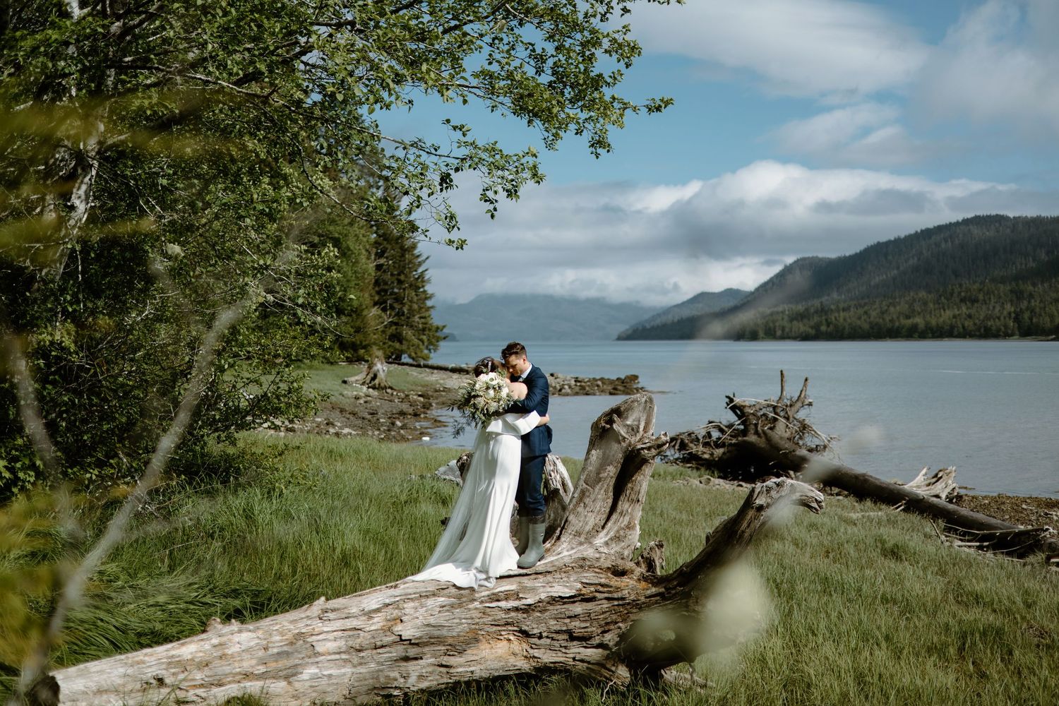 Bride and groom share a romantic kiss on a fallen log by a serene mountain lake surrounded by lush green trees.
