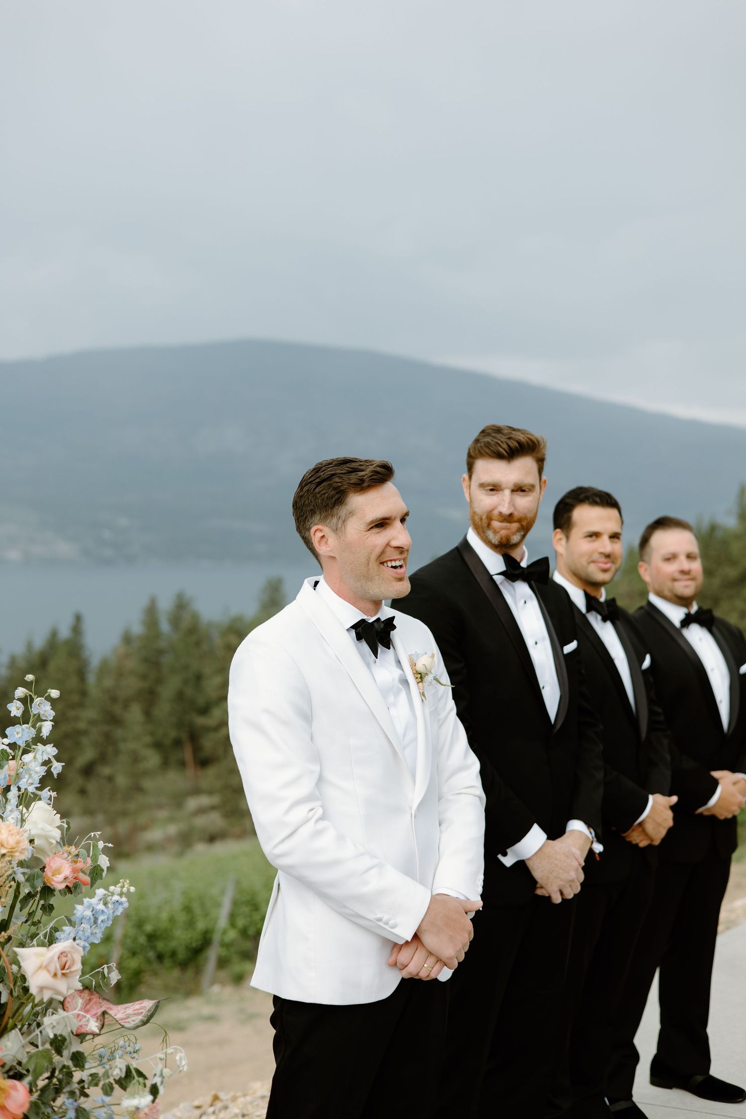 Groomsmen in white tuxedo jackets and black bowties laughing together outdoors.
