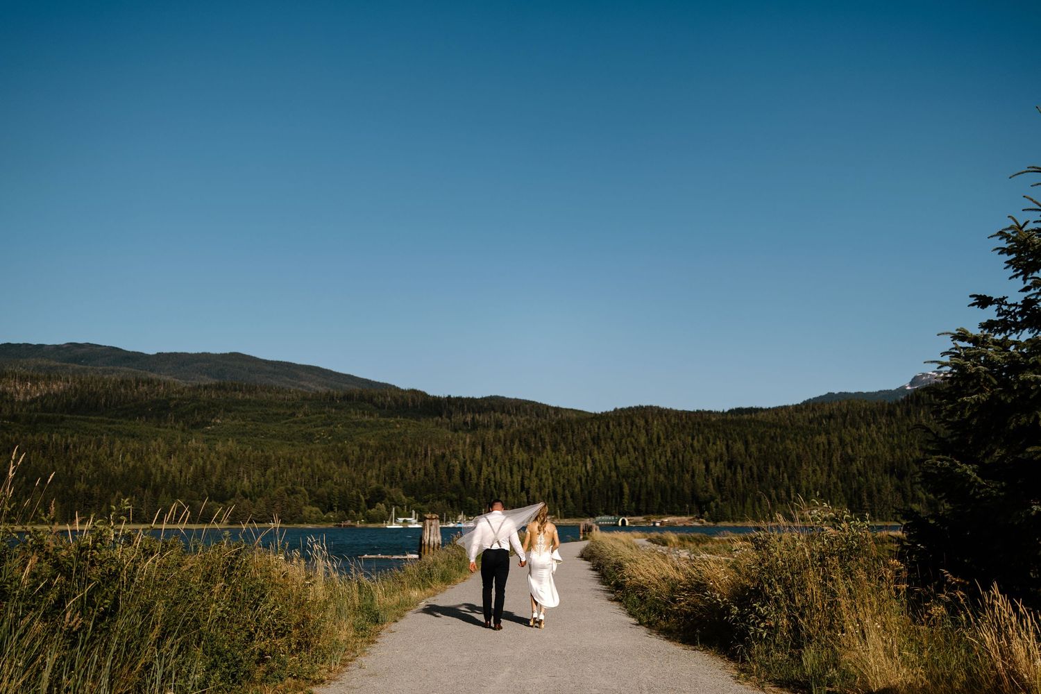 Newlyweds walking down a path with mountain lake and forest scenery in the background.