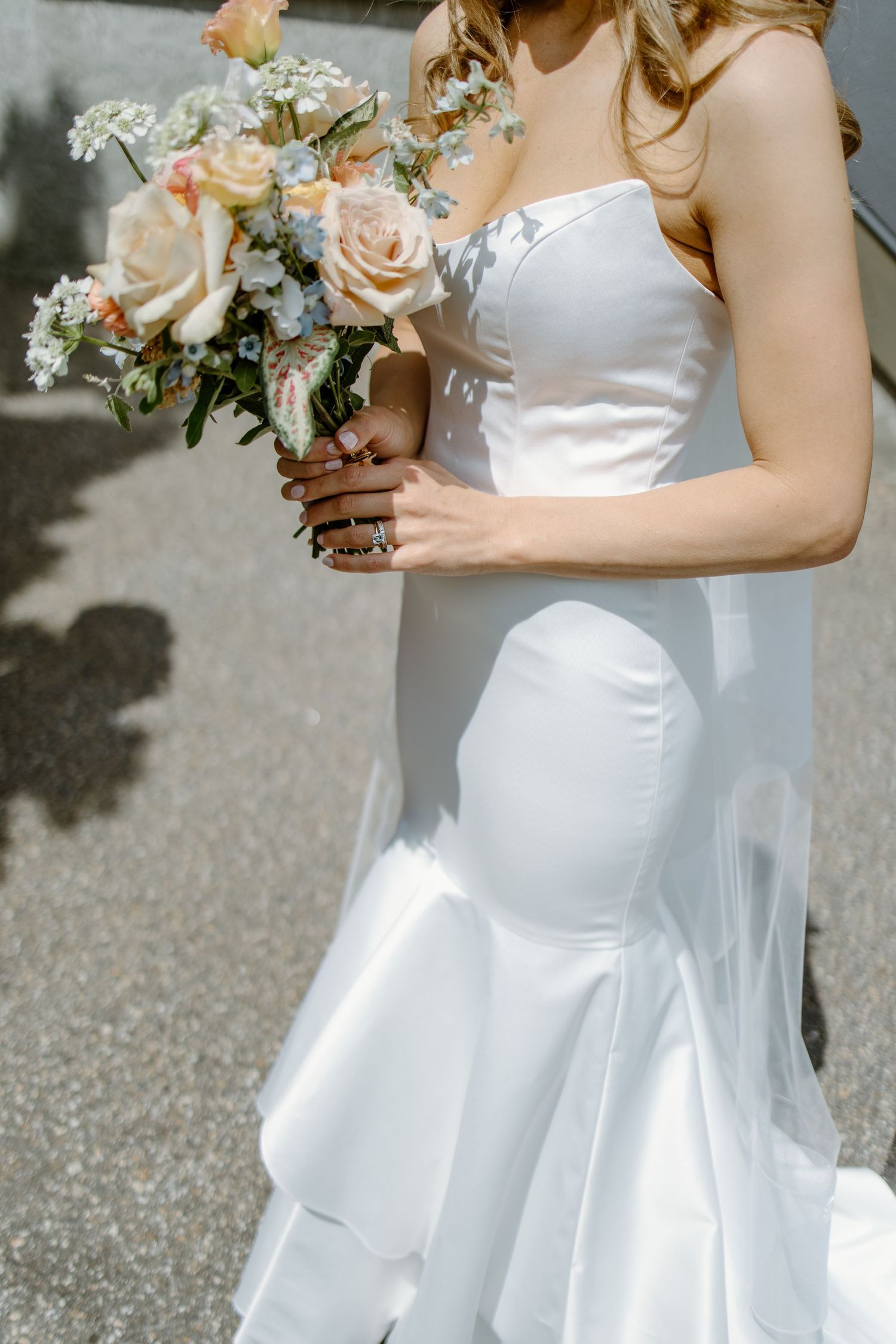 Bride in strapless white mermaid gown holding a pastel bouquet of peach roses and blue flowers outdoors.