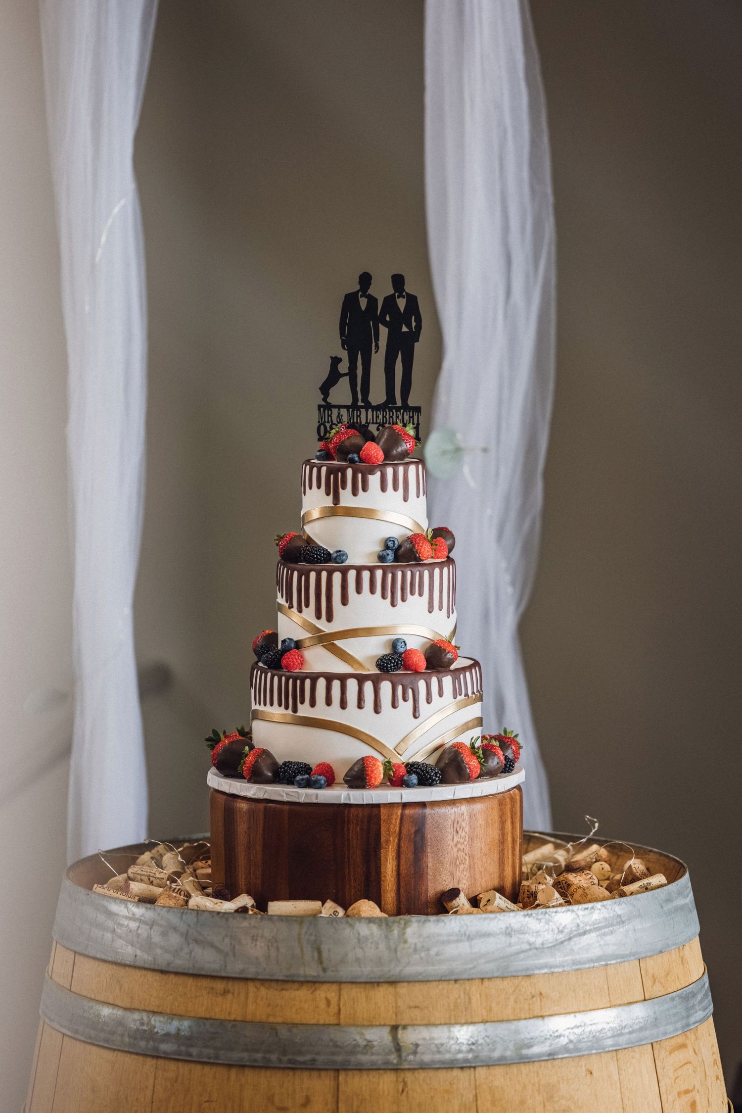 Four-tier wedding cake with berry decorations and same-sex couple topper displayed on a rustic wooden barrel.