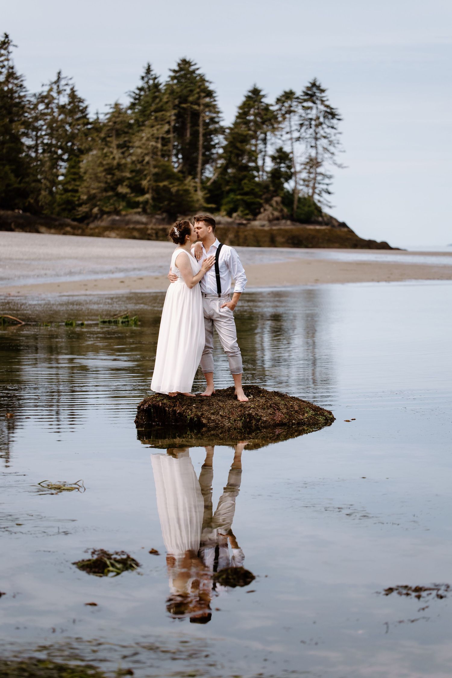 Couple shares intimate moments on a misty beach with evergreen trees and reflective tidal pools at sunset.
