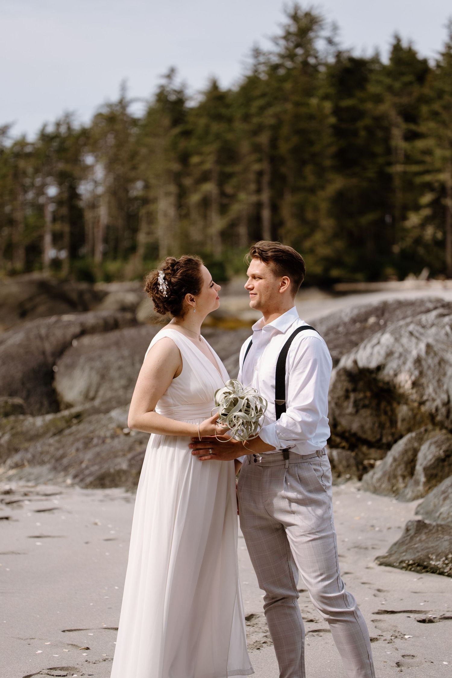 Couple in white exchanging vows on a scenic beach with evergreen trees in background.