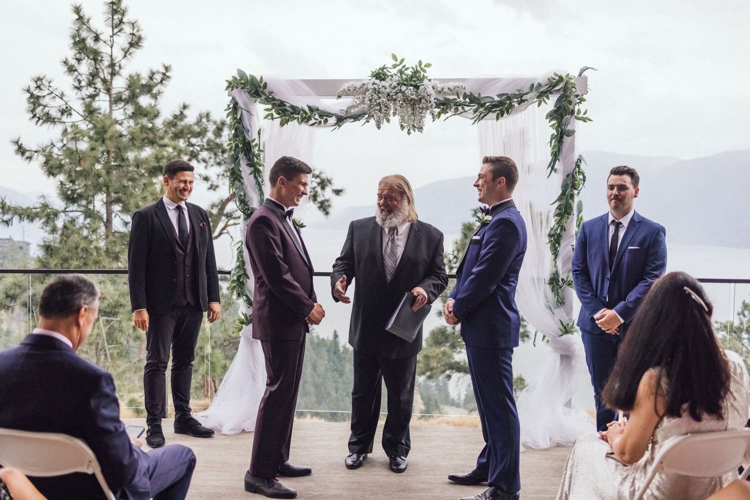 A wedding ceremony takes place under an elegant outdoor arch draped with greenery and white fabric against a mountain backdrop.