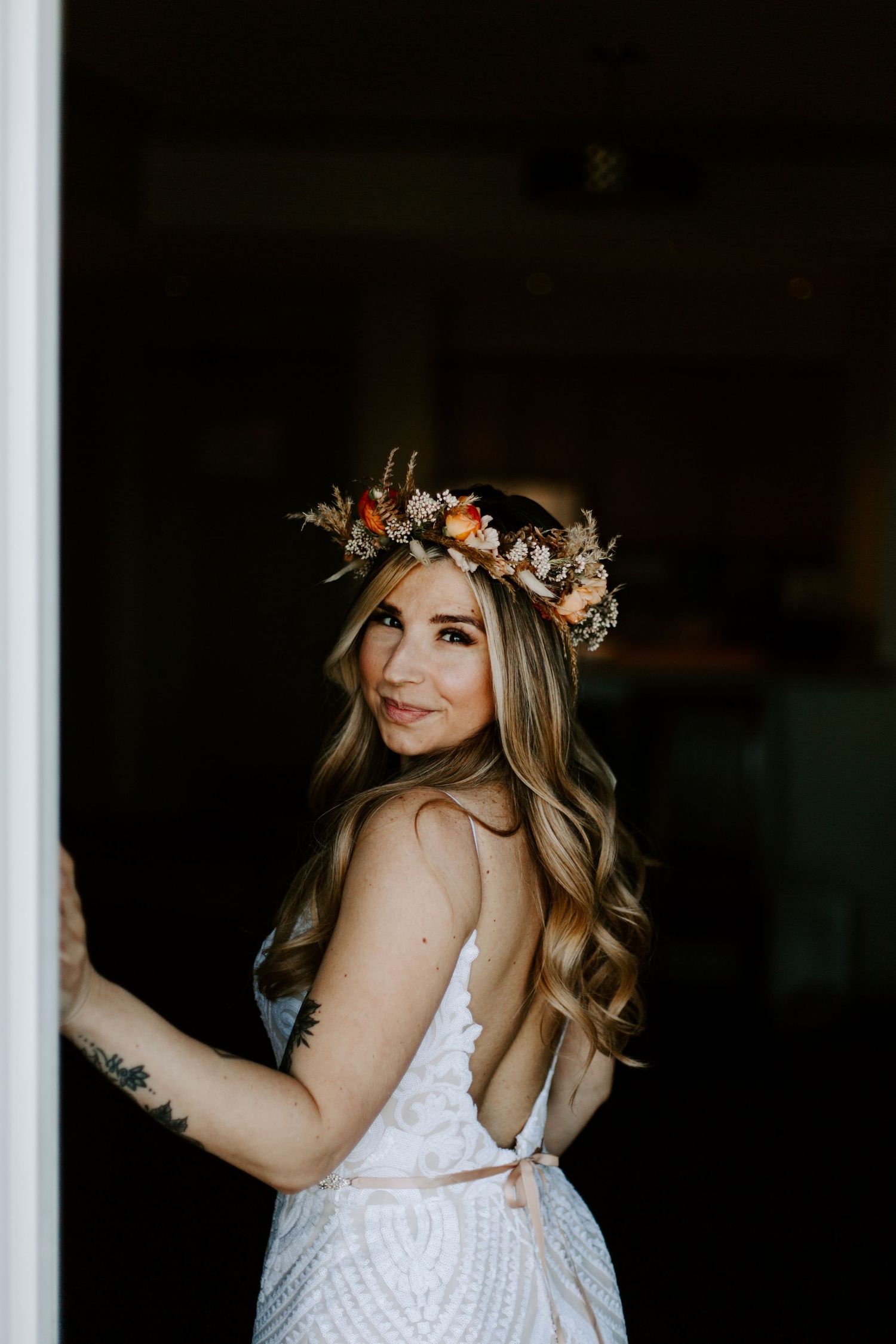 Bride wearing a delicate lace dress and floral crown poses near window light.