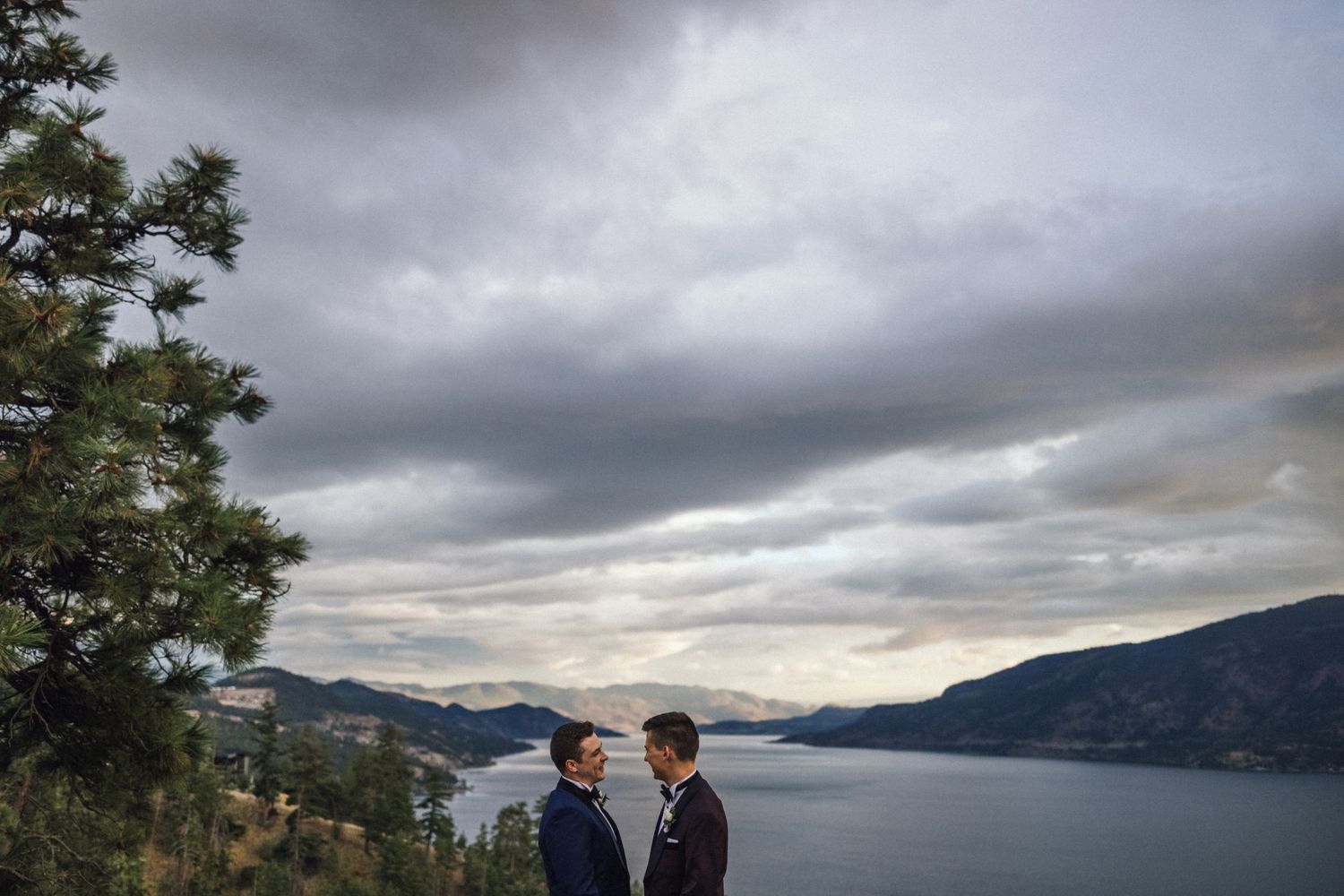 A couple stands silhouetted against dramatic storm clouds and a mountain lake vista.