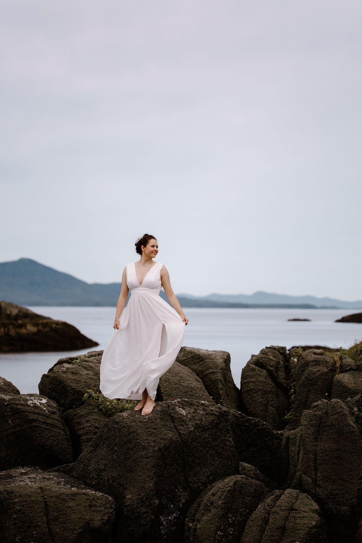 Person in flowing white dress poses gracefully on rocky coastal cliffs overlooking the ocean.