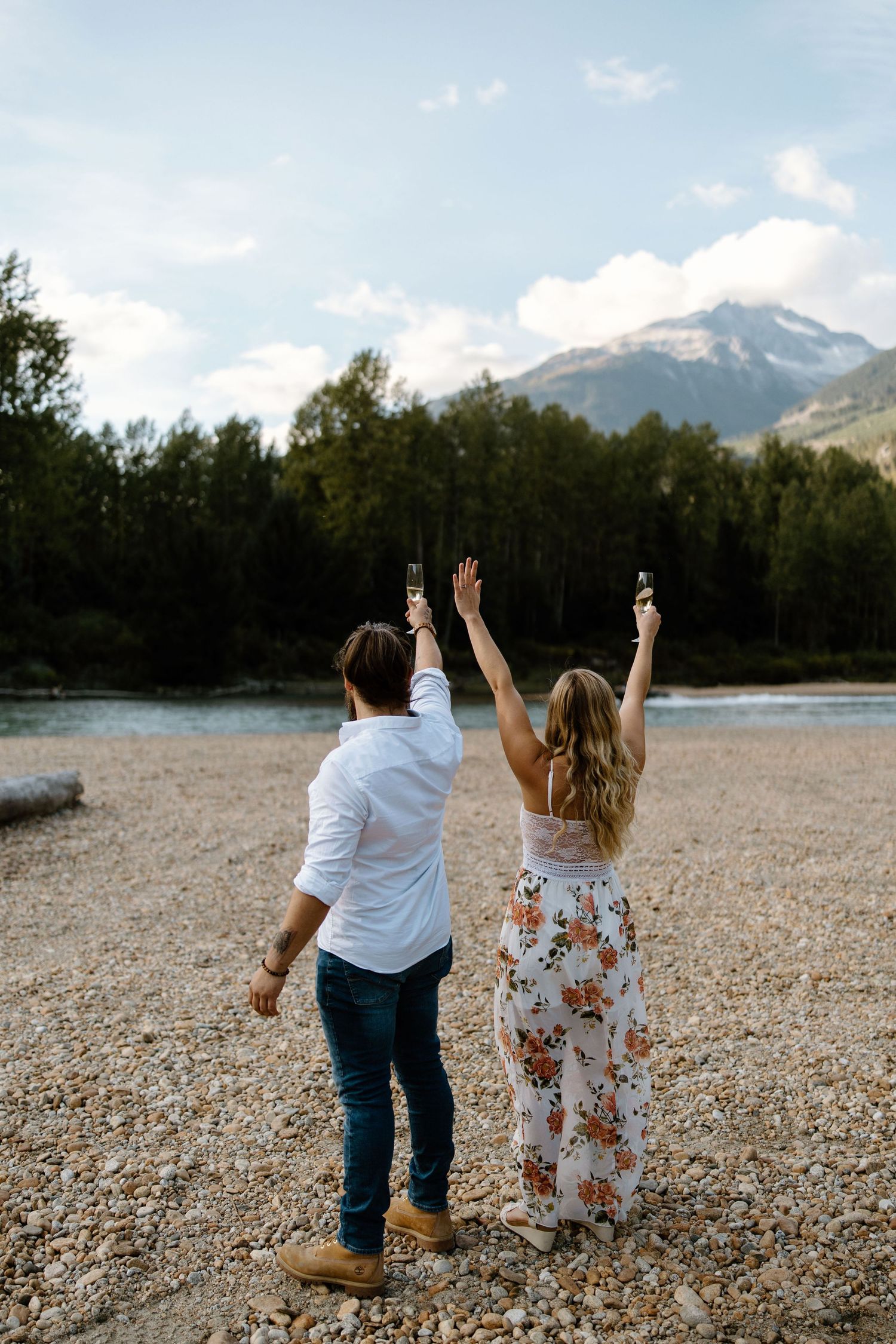 A romantic couple celebrates joyfully on a rocky beach with mountains and forest in the background.