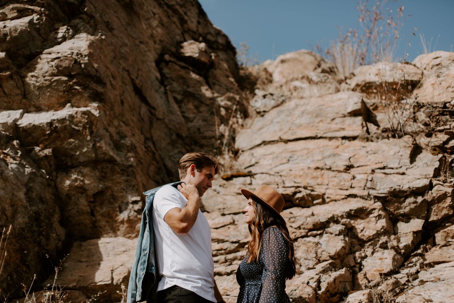 A couple stands close together near rocky cliffs outdoors, the man in a white tee and the woman wearing a brown hat.