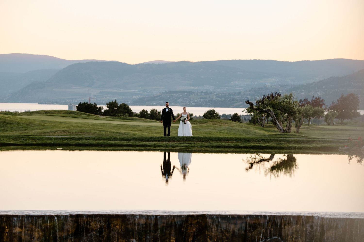 Bride and groom stand by a reflective pond at sunset with scenic lake and mountain views on a golf course.